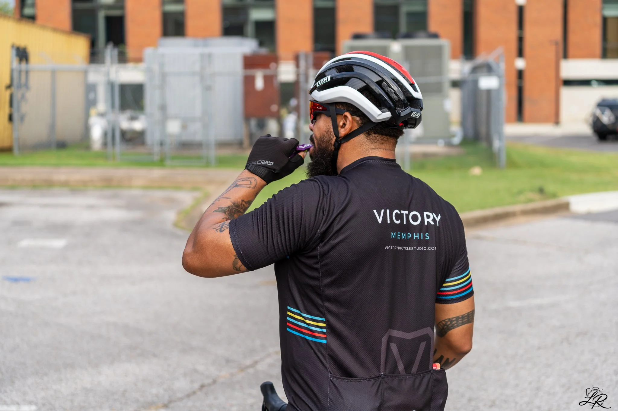 A man wearing a cycling helmet and a black cycling jersey with colorful stripes on the sleeve, standing outdoors near an industrial area, taking a sip from his water bottle.