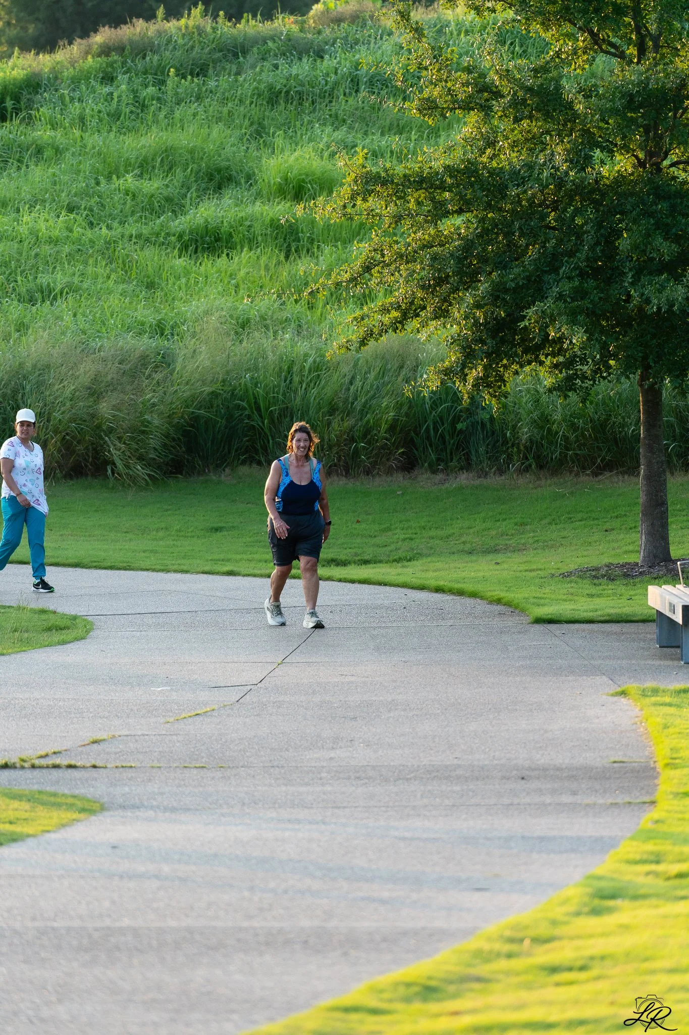 Two women walking on a paved path in a park with green grass and trees, one wearing a white shirt and teal pants with a white cap, and the other in a black skirt and blue tank top.