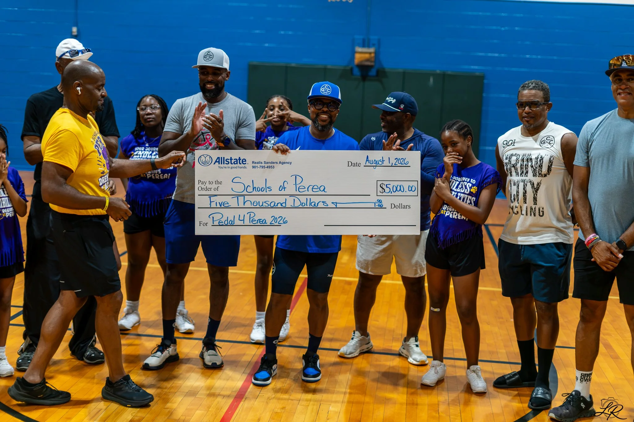 Group of people gathered in a gymnasium, holding a large check for $5,000 made out to Schools of Perea, dated August 1, 2026, surrounded by smiling and clapping individuals, some wearing athletic clothing and hats.