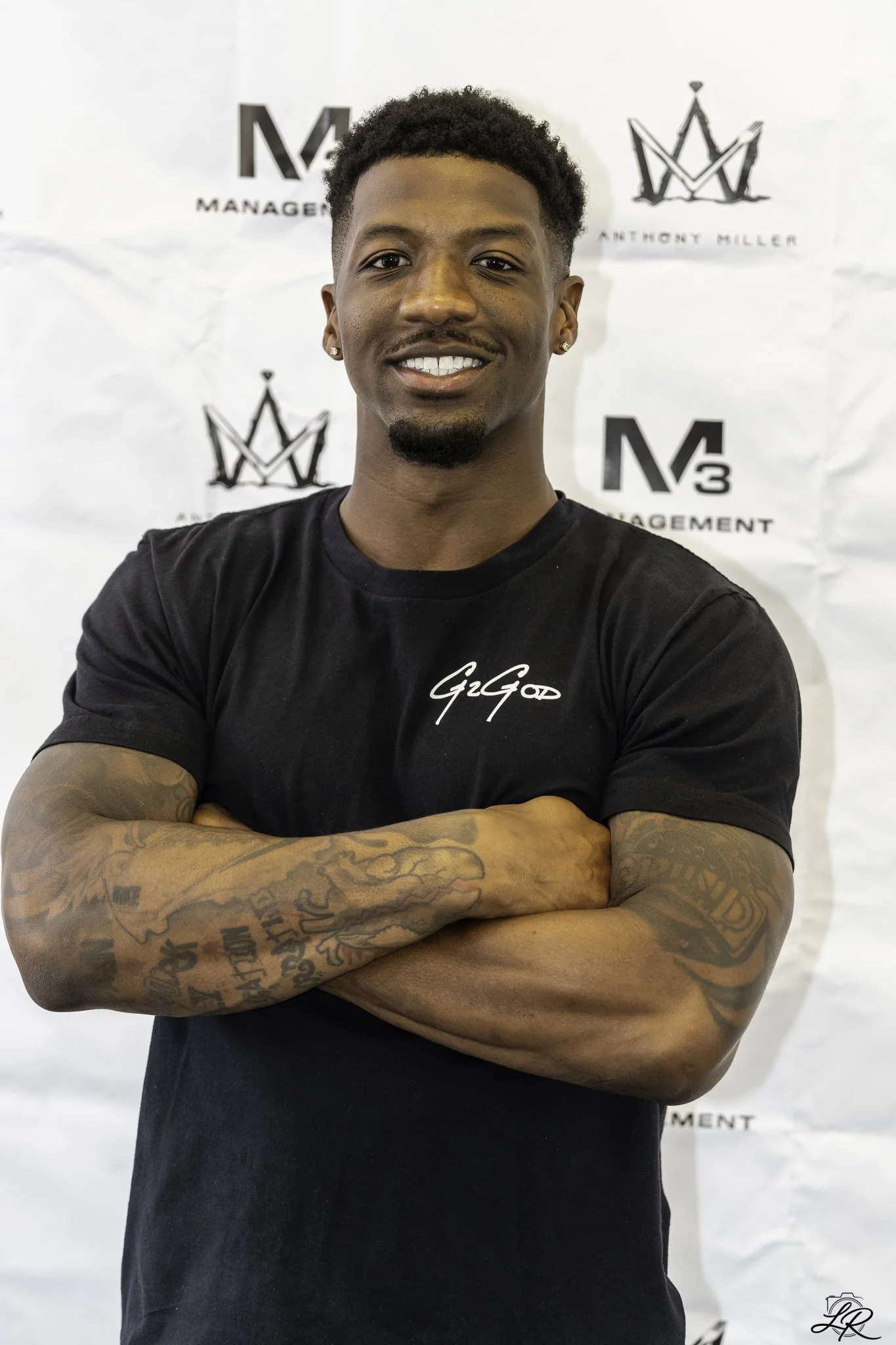 A young man with short curly hair and earrings, smiling and standing with arms crossed in front of a white backdrop with black logos and text.