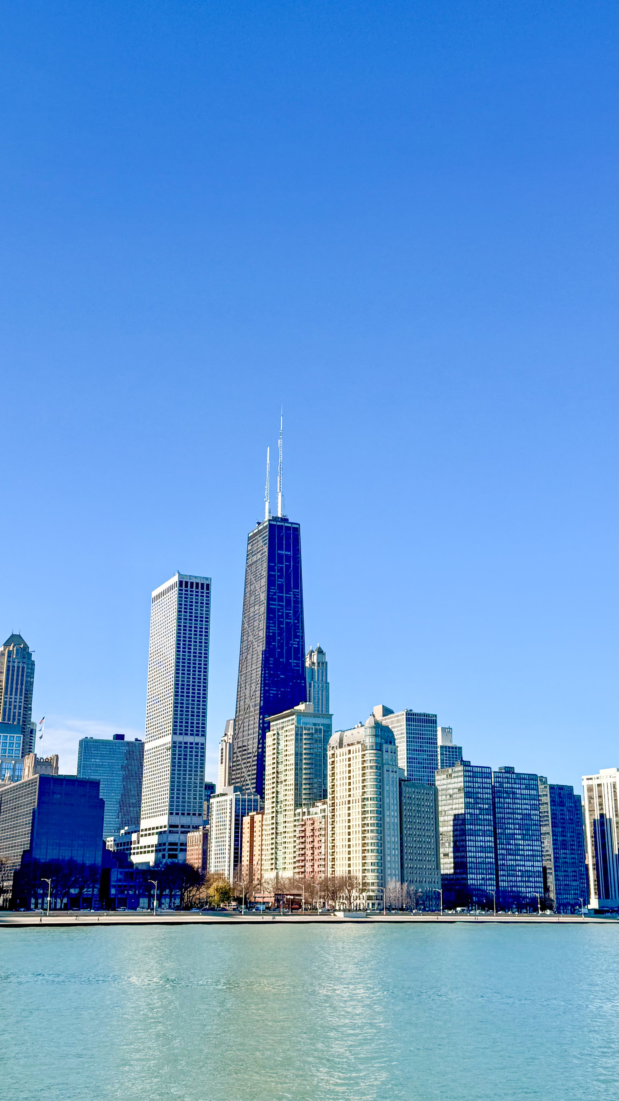 Chicago skyline featuring the John Hancock Center with tall glass buildings reflecting sunlight, seen from across Lake Michigan on a clear day.