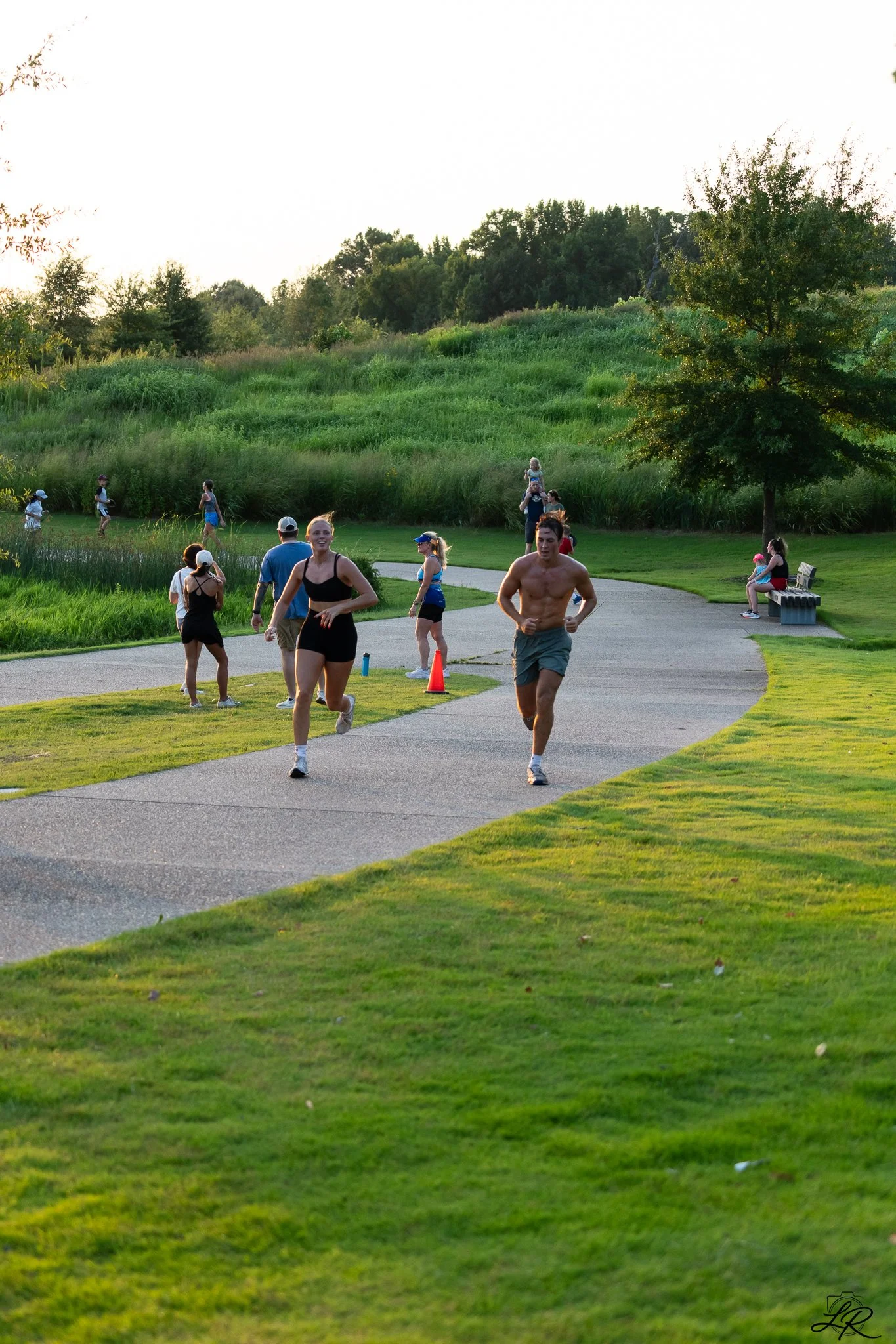 People jogging and walking in a park during sunset, with green grass, trees, and a curved concrete path.