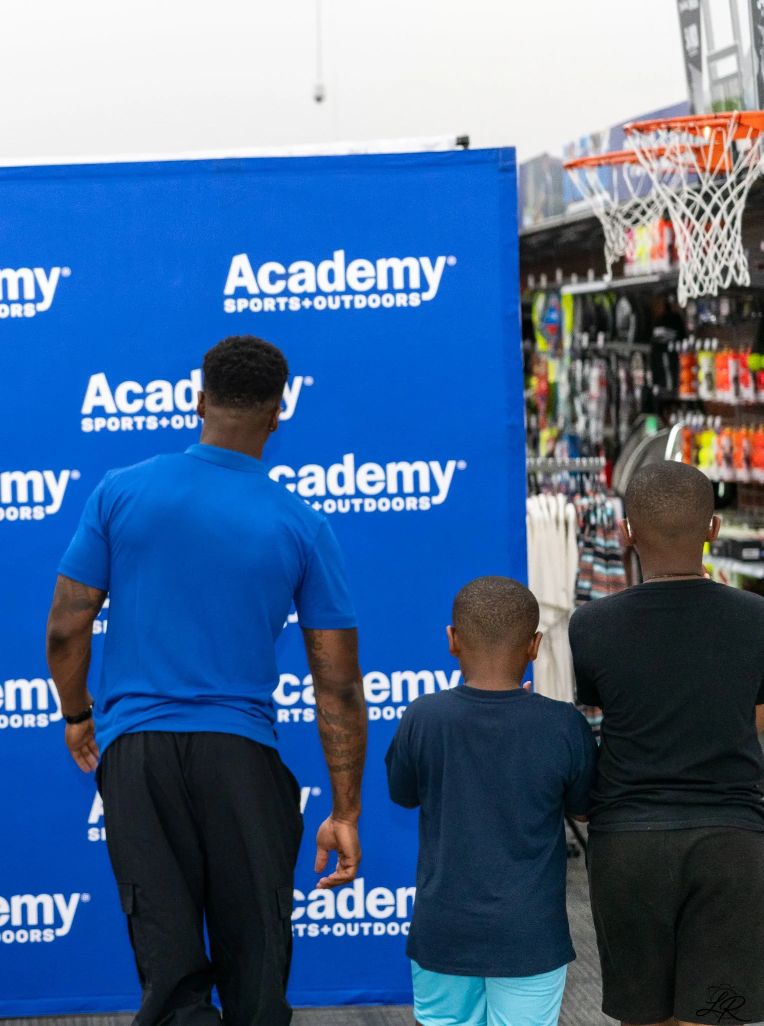 People standing in front of a blue banner with 'Academy SPORTS + OUTDOORS' logo in a store aisle, with a basketball hoop visible to the right.