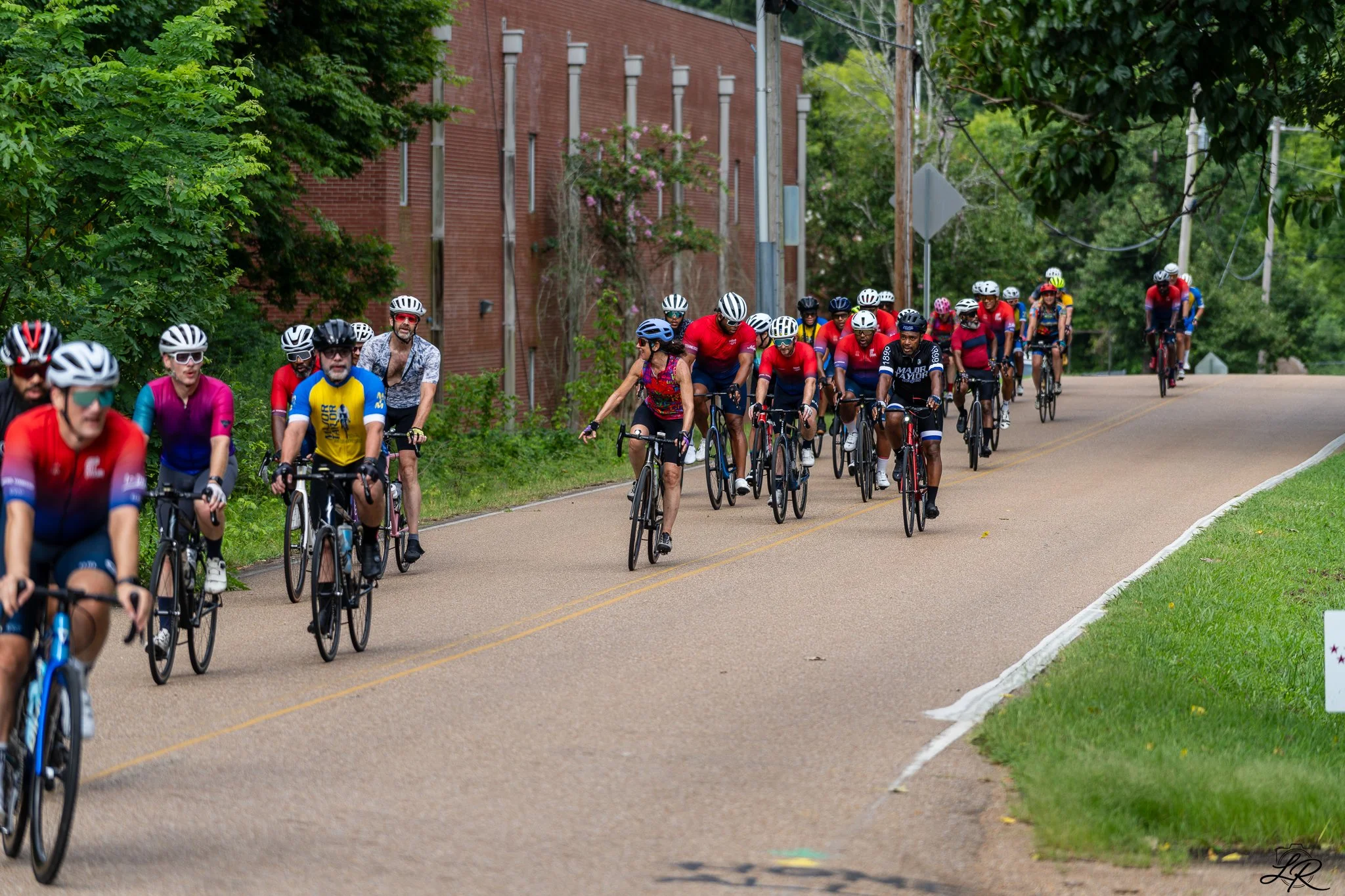 A group of cyclists riding on a paved street surrounded by green trees and a red brick building. The cyclists are wearing helmets and colorful athletic clothing.