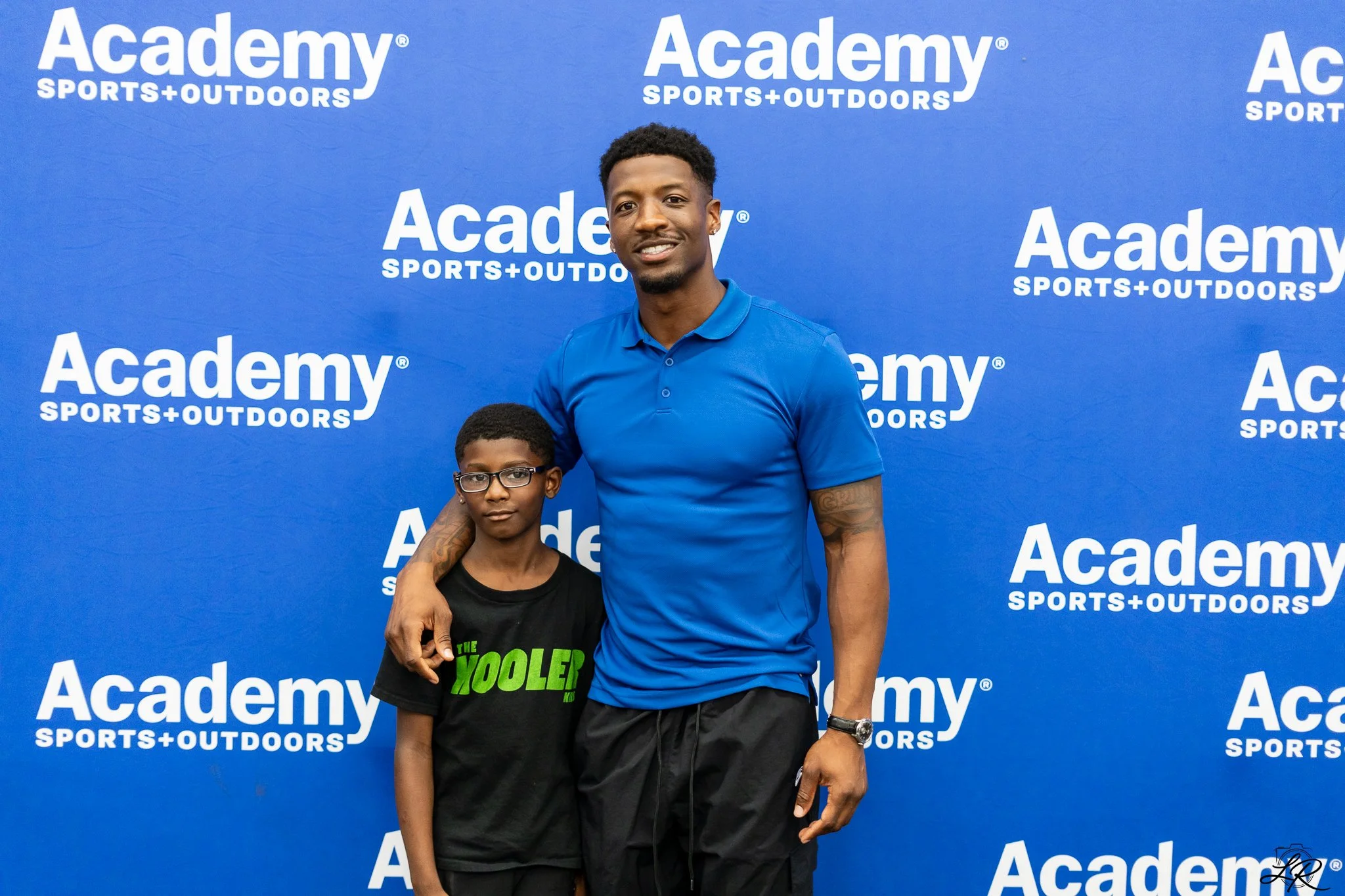 Two males, one young boy and an older man, stand in front of a blue backdrop with the 'Academy Sports + Outdoors' logo repeated across it. The older man is wearing a blue polo shirt and has his arm around the young boy, who is wearing glasses and a b