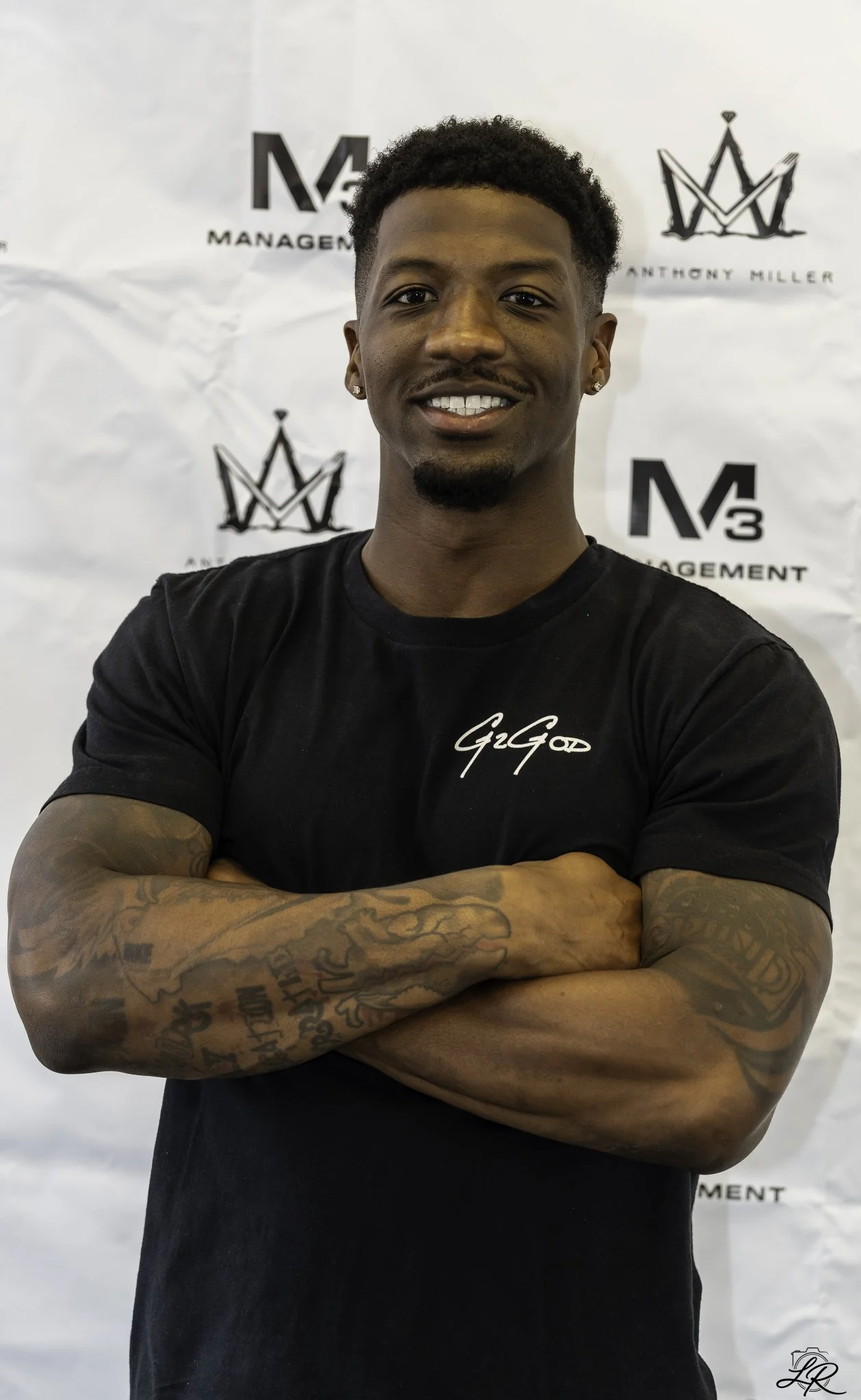 Young man with tattoos on arms, crossing arms, smiling, wearing a black T-shirt with white writing, standing in front of a white background with logos and text.
