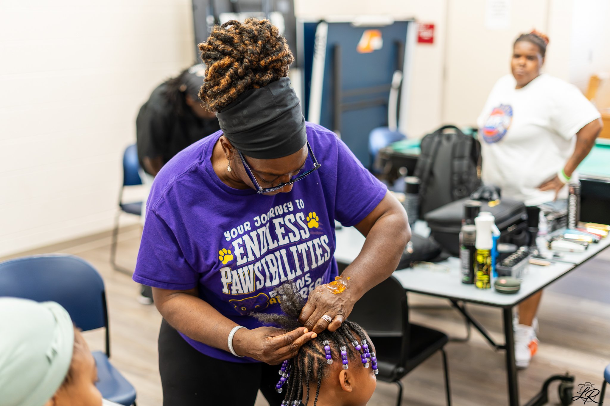 A woman with curly hair standing and working on a young girl with braids decorated with purple beads in a room with tables, chairs, and various hair products.