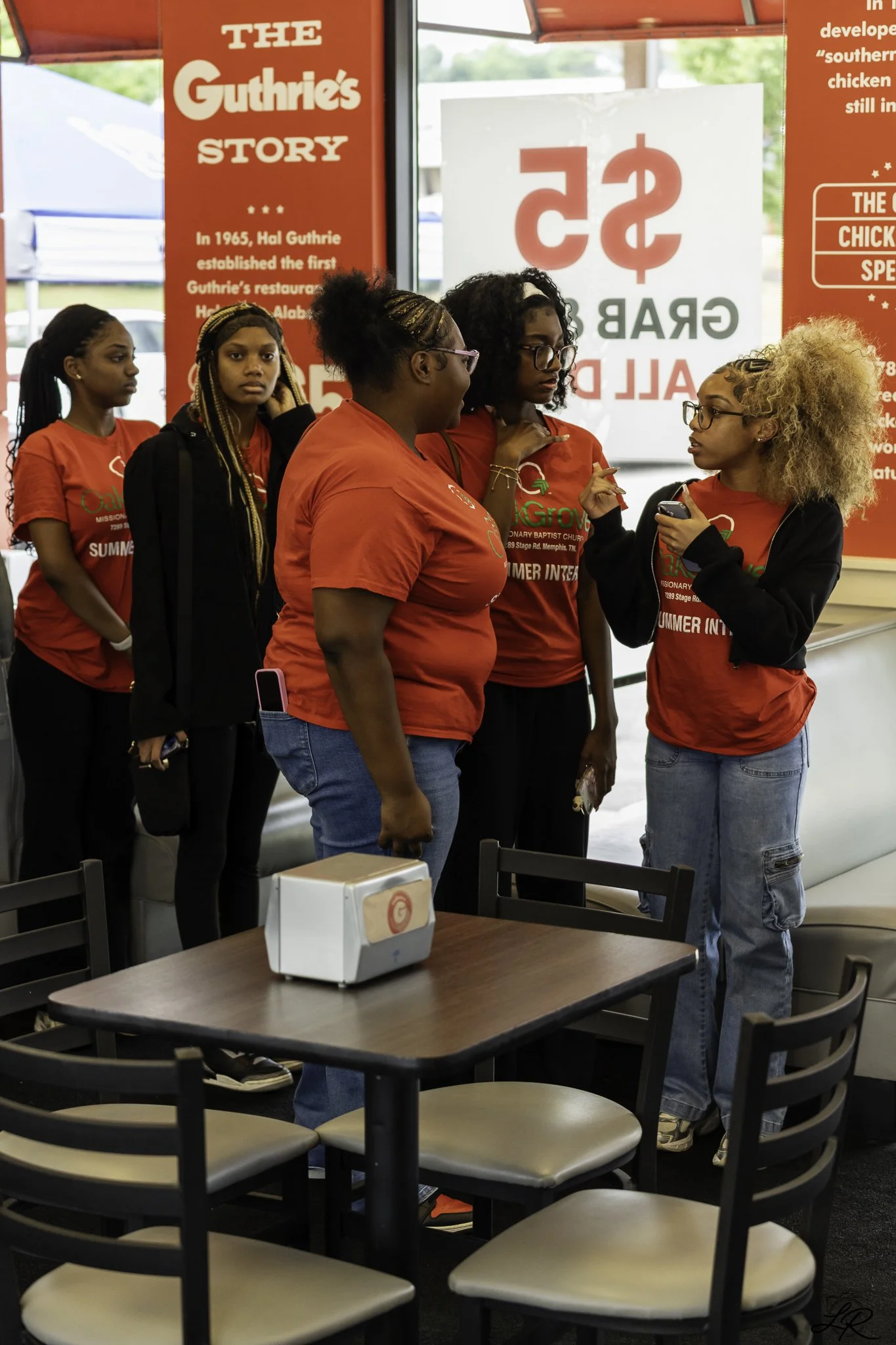 A group of people standing together inside a restaurant, wearing red shirts with green and white logos, engaged in a conversation. The restaurant has red promotional posters on the windows and empty tables and chairs in the foreground.