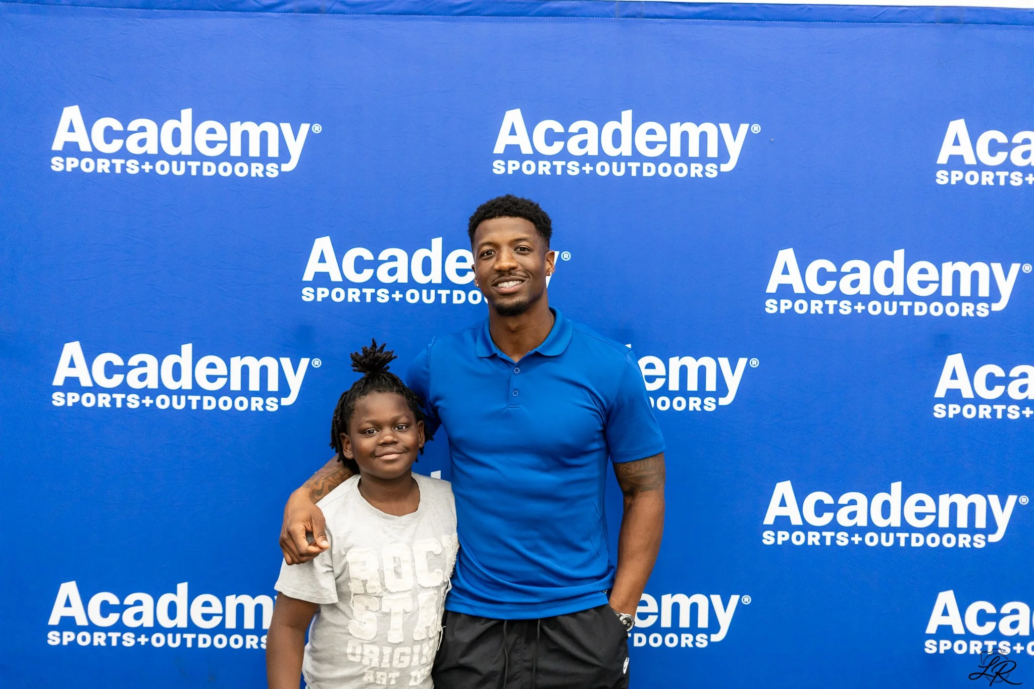 A young man with short curly hair wearing a blue polo shirt and black shorts is smiling and standing with his arm around a young girl with braided hair and a light-colored T-shirt. They are standing in front of a blue backdrop with the white text 'Ac