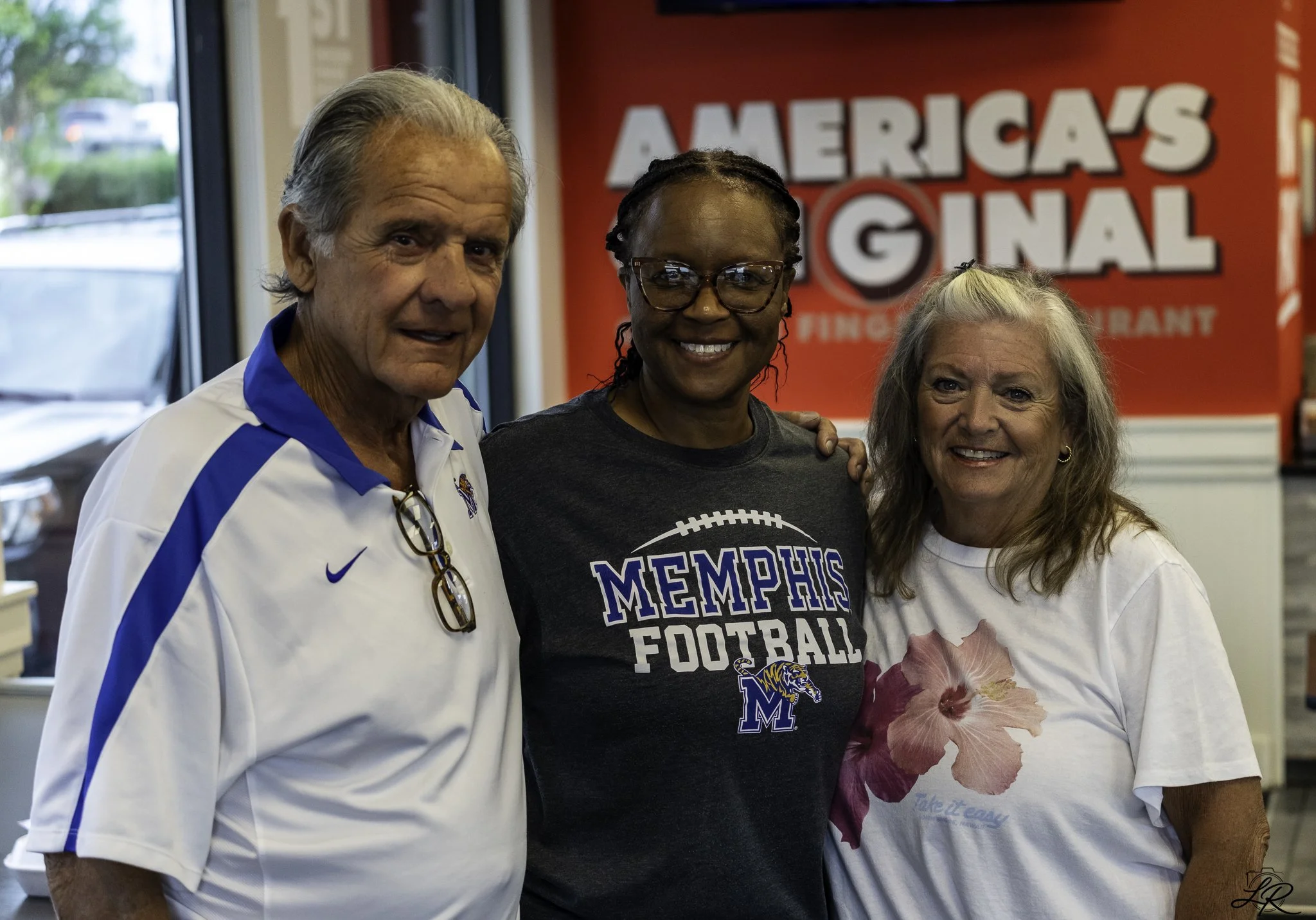 Three smiling women standing in front of a red sign that reads 'AMERICAS' and 'NATIONAL'. One woman is wearing a Memphis football t-shirt, and another is holding a pink flower and wearing a white t-shirt with a floral design.
