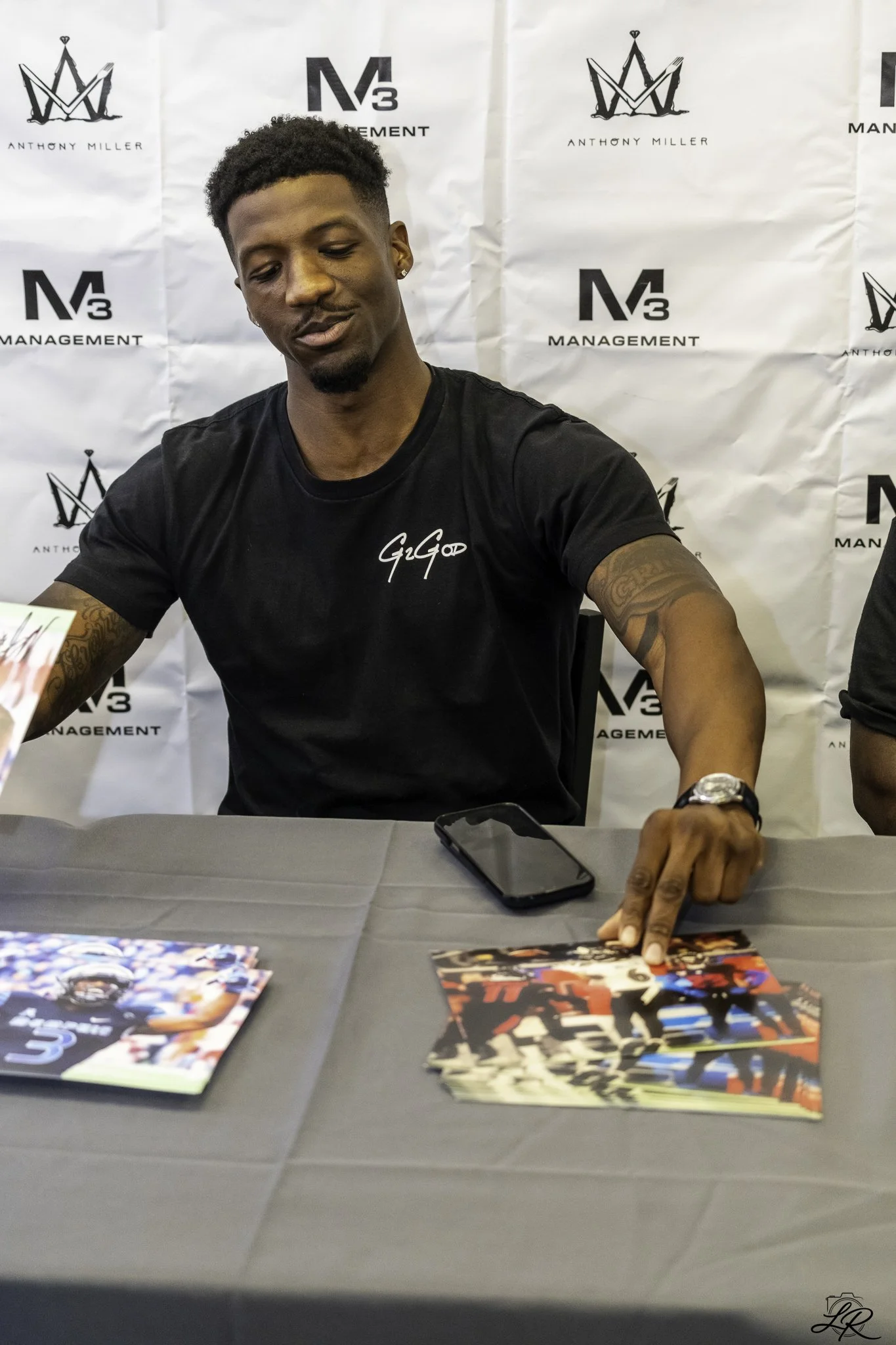 A young man with short curly hair and earrings, wearing a black shirt with white script on it, sitting at a table with photographs and posters, pointing to one of the posters. The background features a step-and-repeat banner with logos and the text A