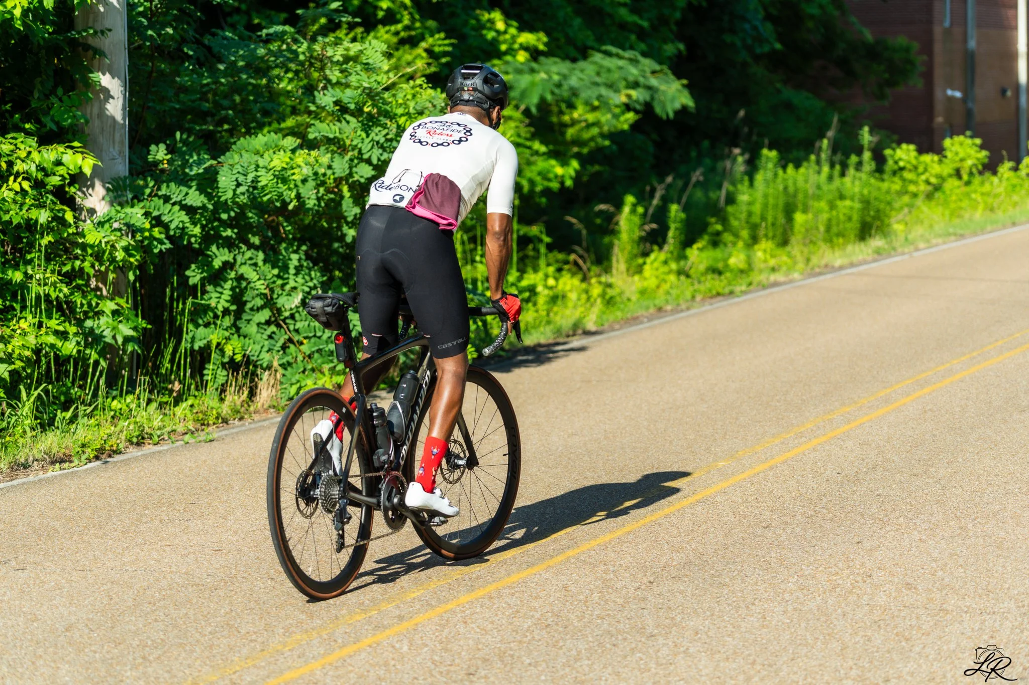 A cyclist wearing a black helmet and white jersey riding a black road bike on a paved road with yellow lines, surrounded by green foliage and trees on a sunny day.