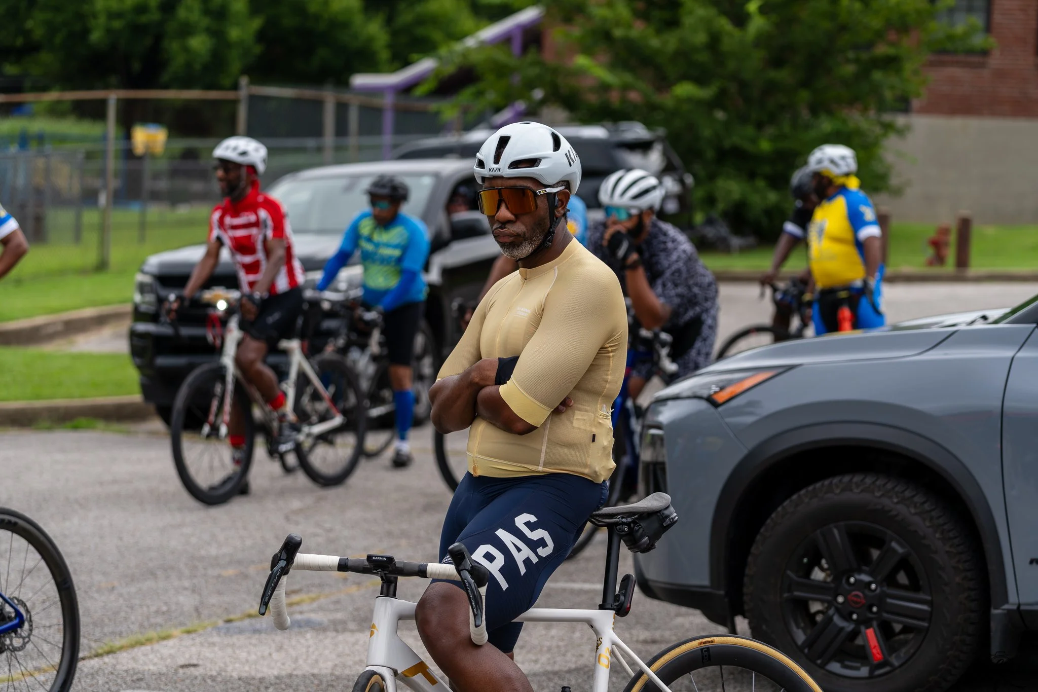 A man wearing a white cycling helmet, sunglasses, a tan long-sleeve shirt, and dark shorts with the word 'PAS' visible, standing with folded arms near his bicycle during a group cycling event. In the background, other cyclists in colorful gear are pr
