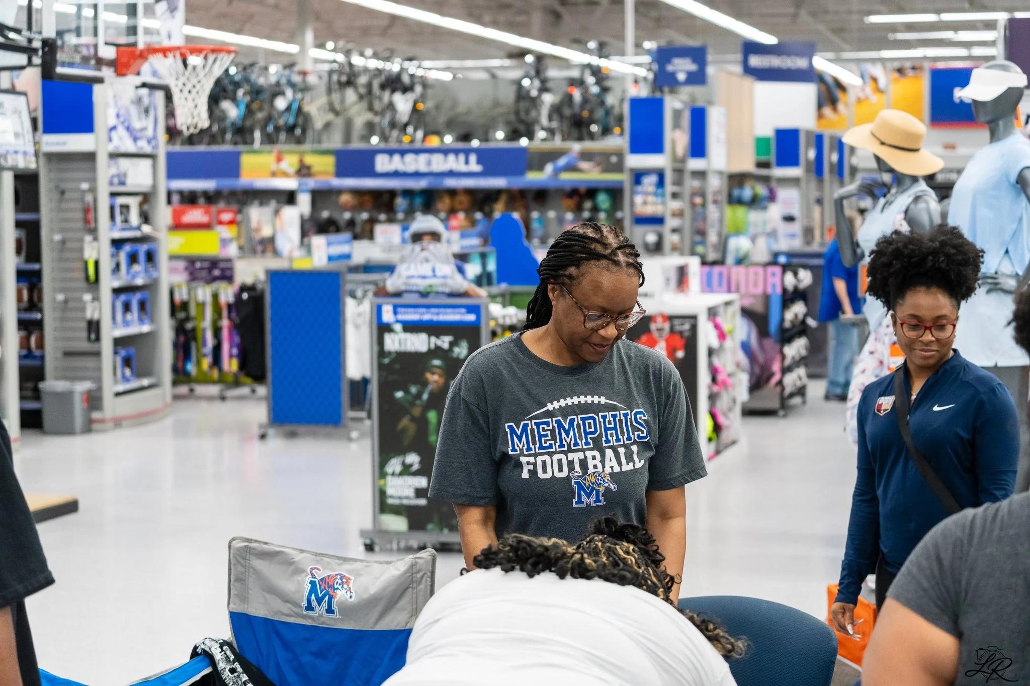 Two women in Memphis football shirts shopping in a big retail store with sports and outdoor gear. One woman with glasses and braids is looking down, and the other woman with glasses and curly hair is smiling. Various sports items and displays are in 