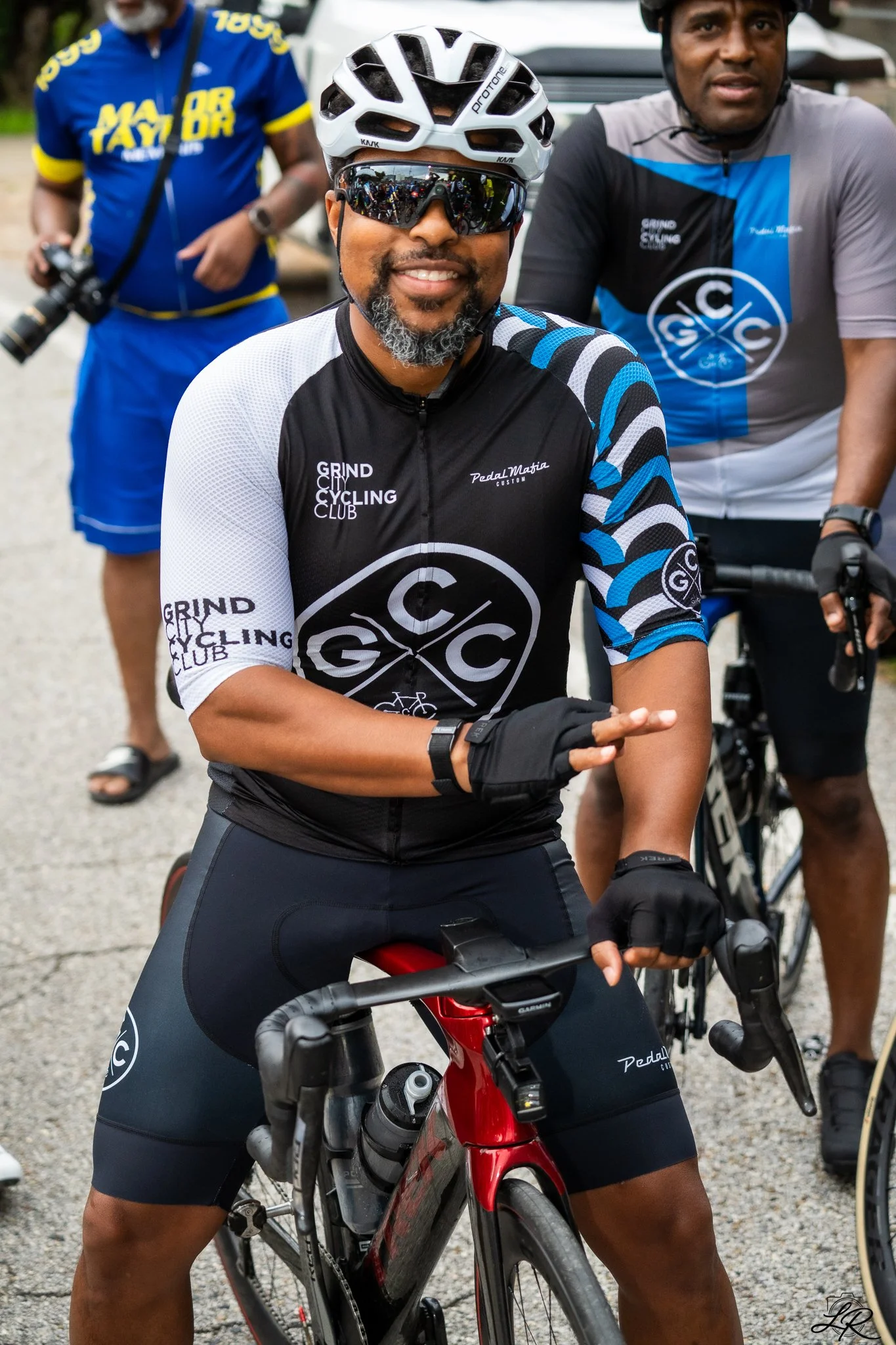 Three men posing with bicycles, wearing cycling gear and helmets, outdoors on a gravel surface.