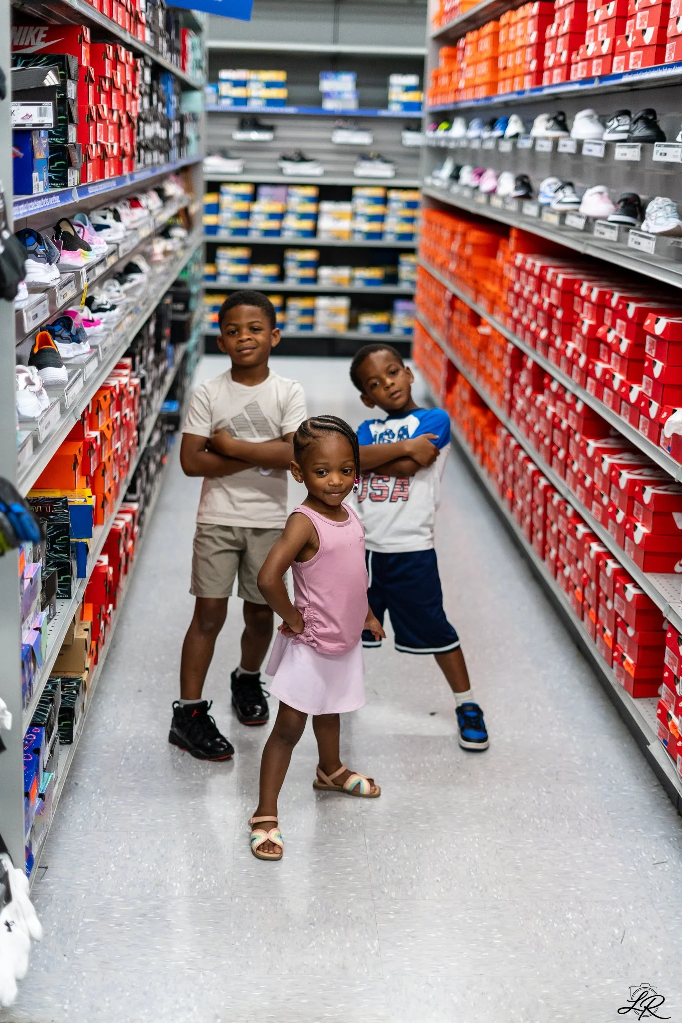 Three children standing in sneaker aisle of a shoe store, with shelves of sneakers and shoe boxes behind them.