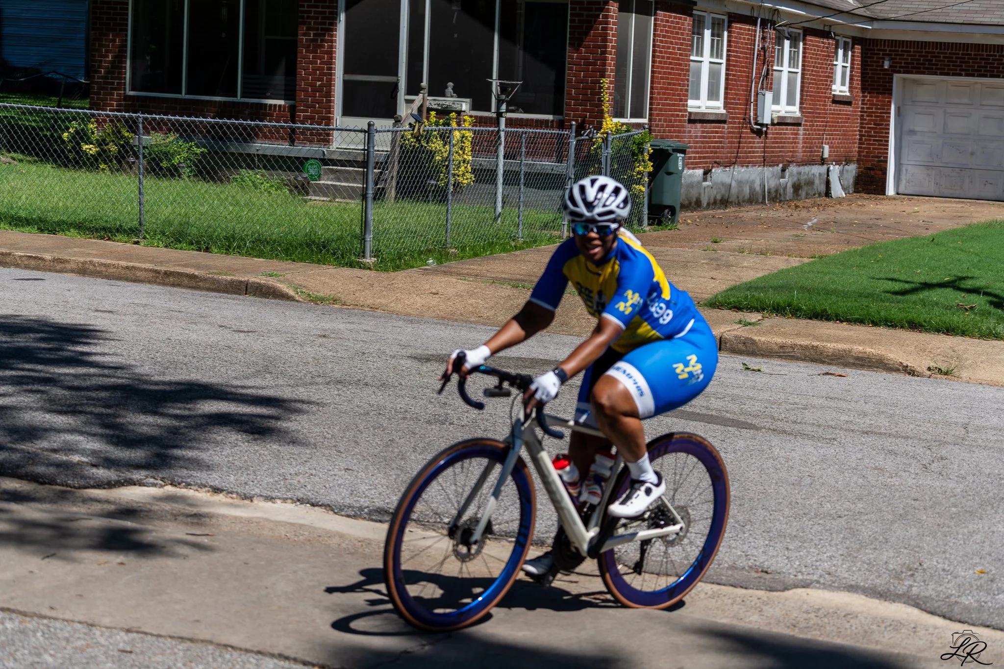 Child wearing a helmet and sunglasses riding a bike on the street with a brick house and green lawn in the background.