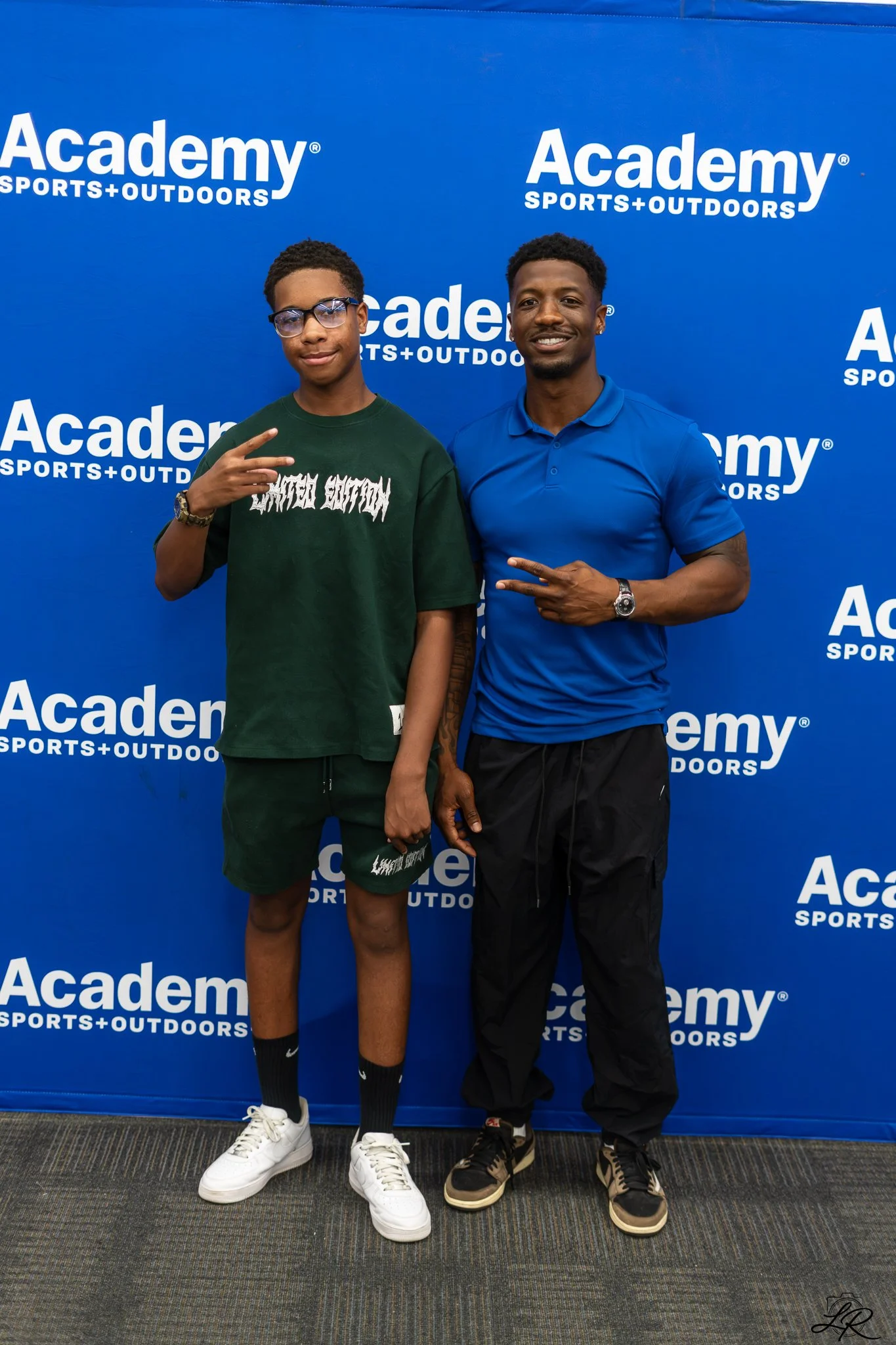 Two men stand side by side against a blue backdrop with 'Academy Sports + Outdoors' written repeatedly. The man on the left is wearing glasses, a green t-shirt with white text, dark shorts, and white sneakers. He is making a peace sign with his right