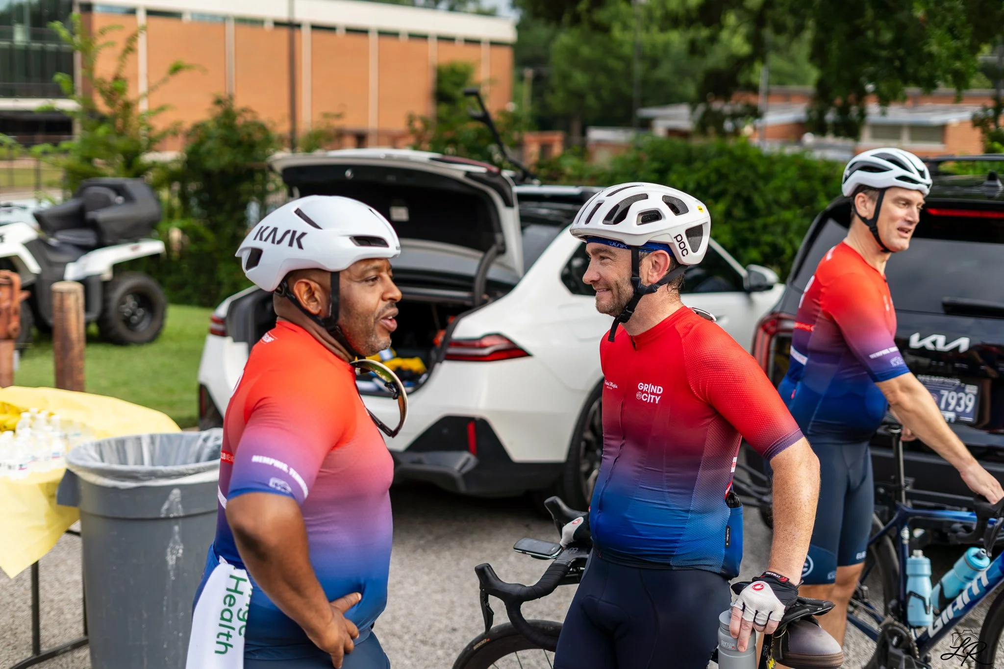 Three cyclists in red into blue gradient cycling jerseys and white helmets standing among parked bikes and cars, engaged in conversation outdoors during daytime.