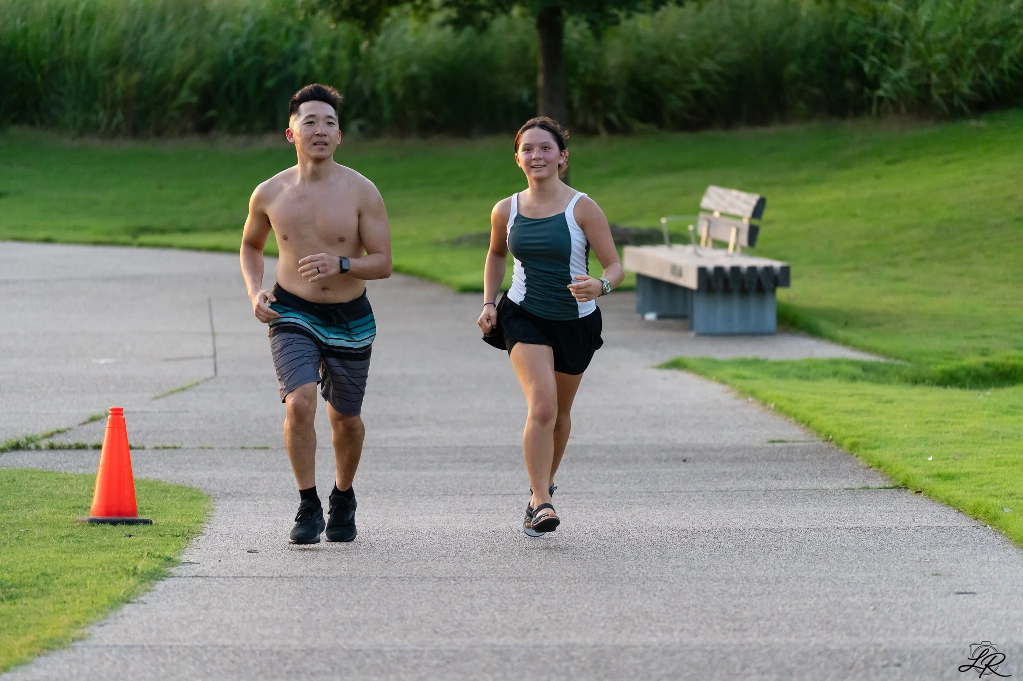 Two people jogging in a park along a paved pathway, with an orange traffic cone nearby and green grass on either side, during daytime.