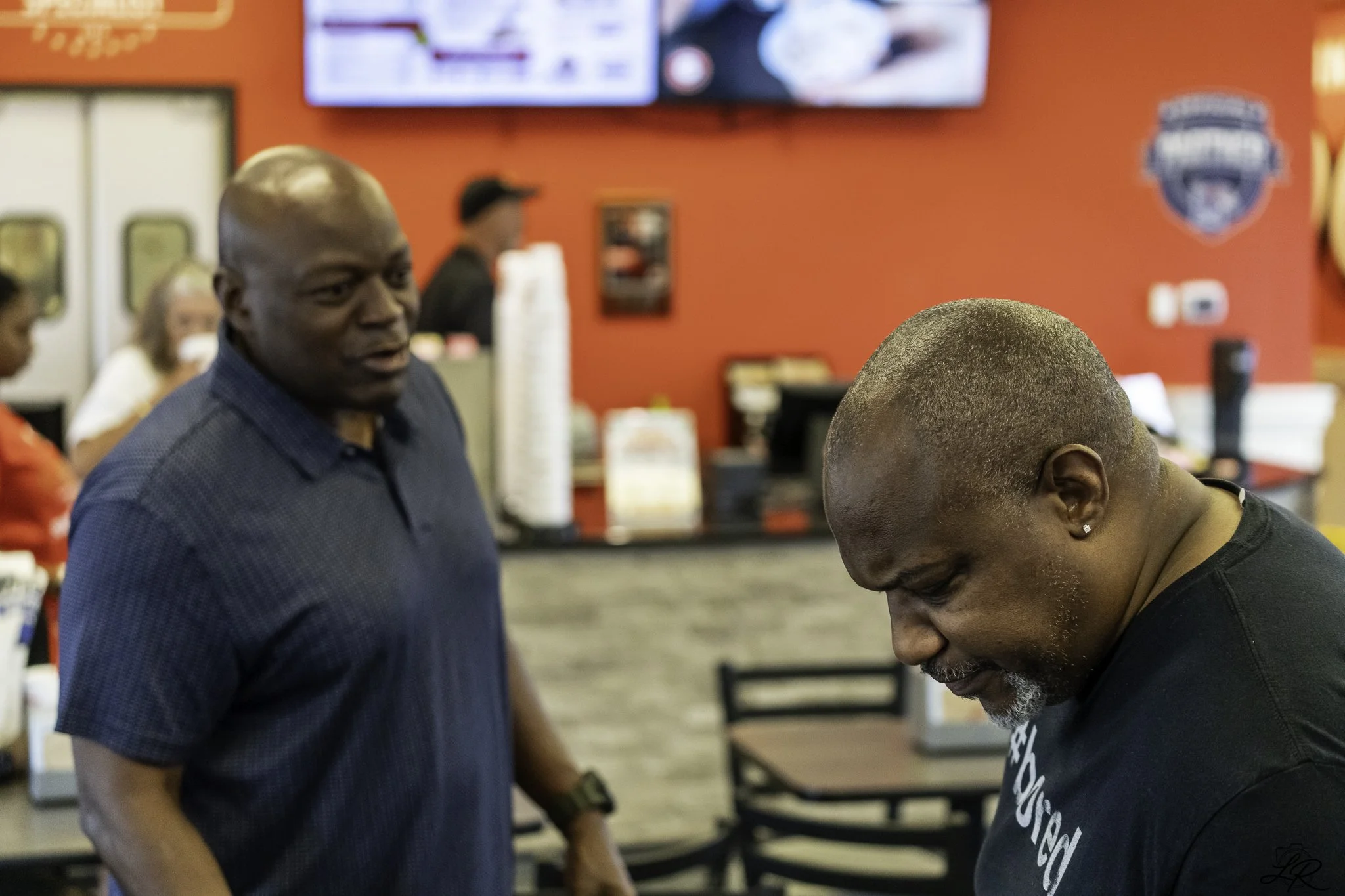 Two men are in a fast-food restaurant. One man with a bald head and a dark shirt is looking at another man with short hair and a beard, who is leaning forward with a smile. The background is red with a flat-screen TV and restaurant branding.