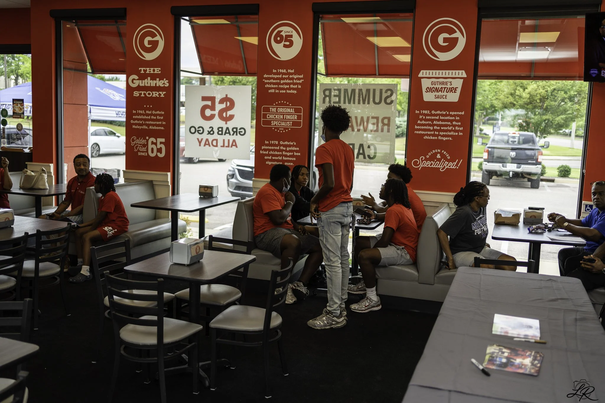 Inside a fast food restaurant with red walls and large windows. Several employees and customers are seated and standing, some wearing masks. There are signs on the windows describing the restaurant's history and signature sauces.
