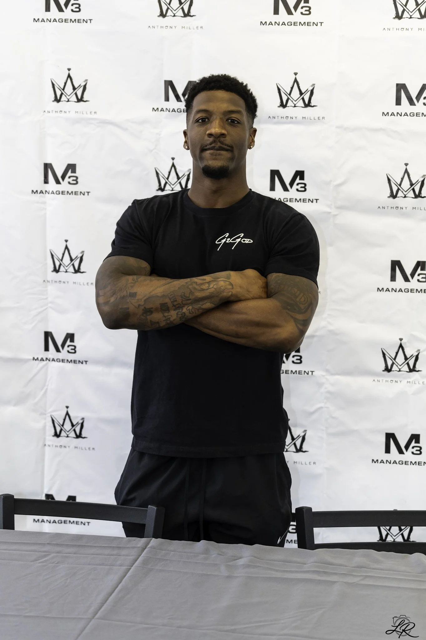 A young man with short curly hair and earrings stands with arms crossed in front of a backdrop marked with logos for N3 Management and Anthony Miller. He is wearing a black T-shirt with white writing on it and black shorts, posing confidently at an e