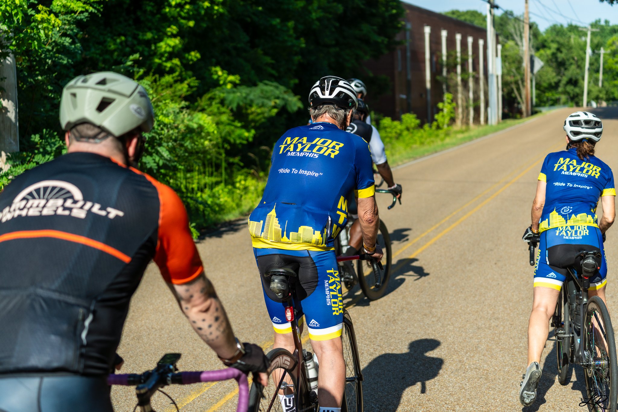 Group of cyclists riding on a rural road with green trees and power lines in the background. Some riders wear blue and yellow jerseys with the text 'Major Taylor Memphis' and 'Ride To Inspire.'