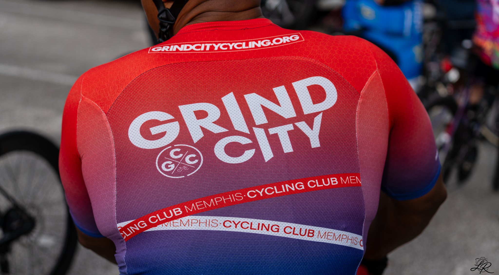 Back of a cyclist wearing a red jersey with 'Grand City' and 'Memphis Cycling Club' printed on it, along with a logo, on a road with bicycles parked in the background.