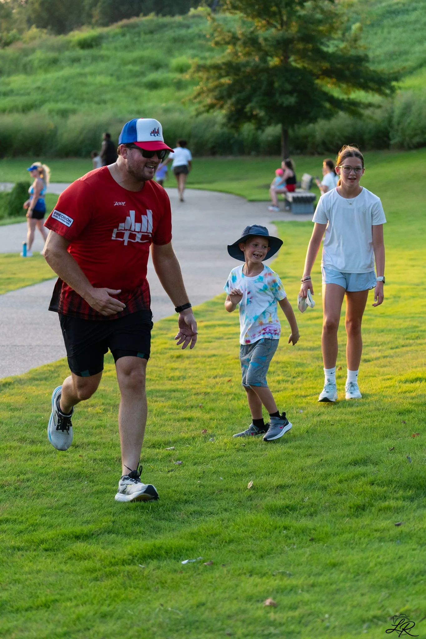 A man and two kids running and walking in a park during the daytime, with a bright green grassy area, trees, and other people in the background.