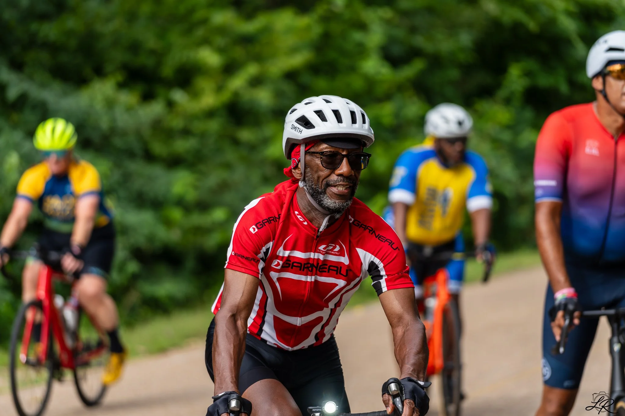 A group of people riding bicycles outdoors with greenery in the background. The man in the foreground is wearing a white helmet, sunglasses, a red cycling jersey, and black shorts.
