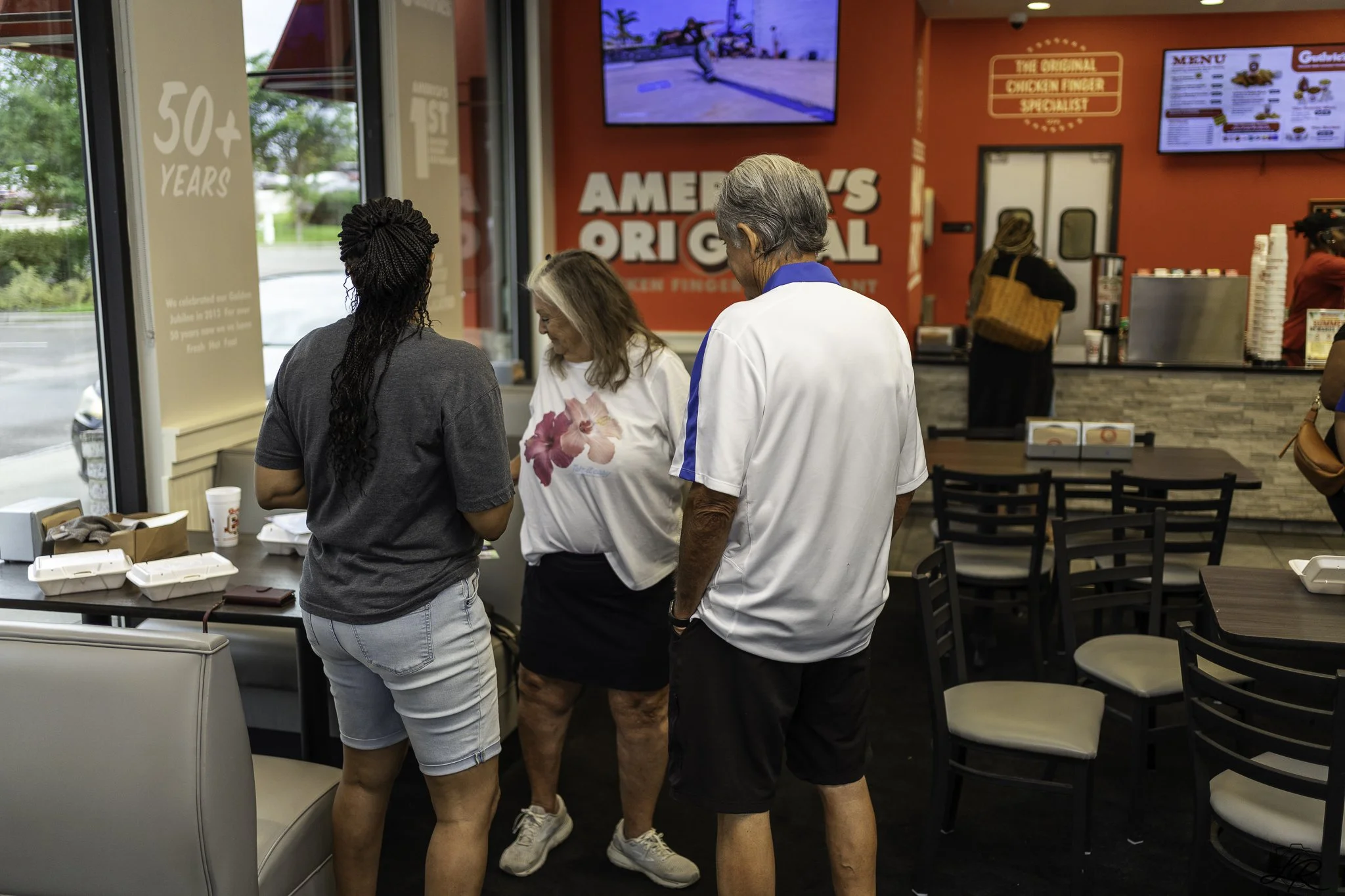 Four people in line inside a fast-food restaurant, with a red interior and a television mounted on the wall. The restaurant has a large sign that reads 'America's Original.' The group includes three women and one man, all casually dressed.