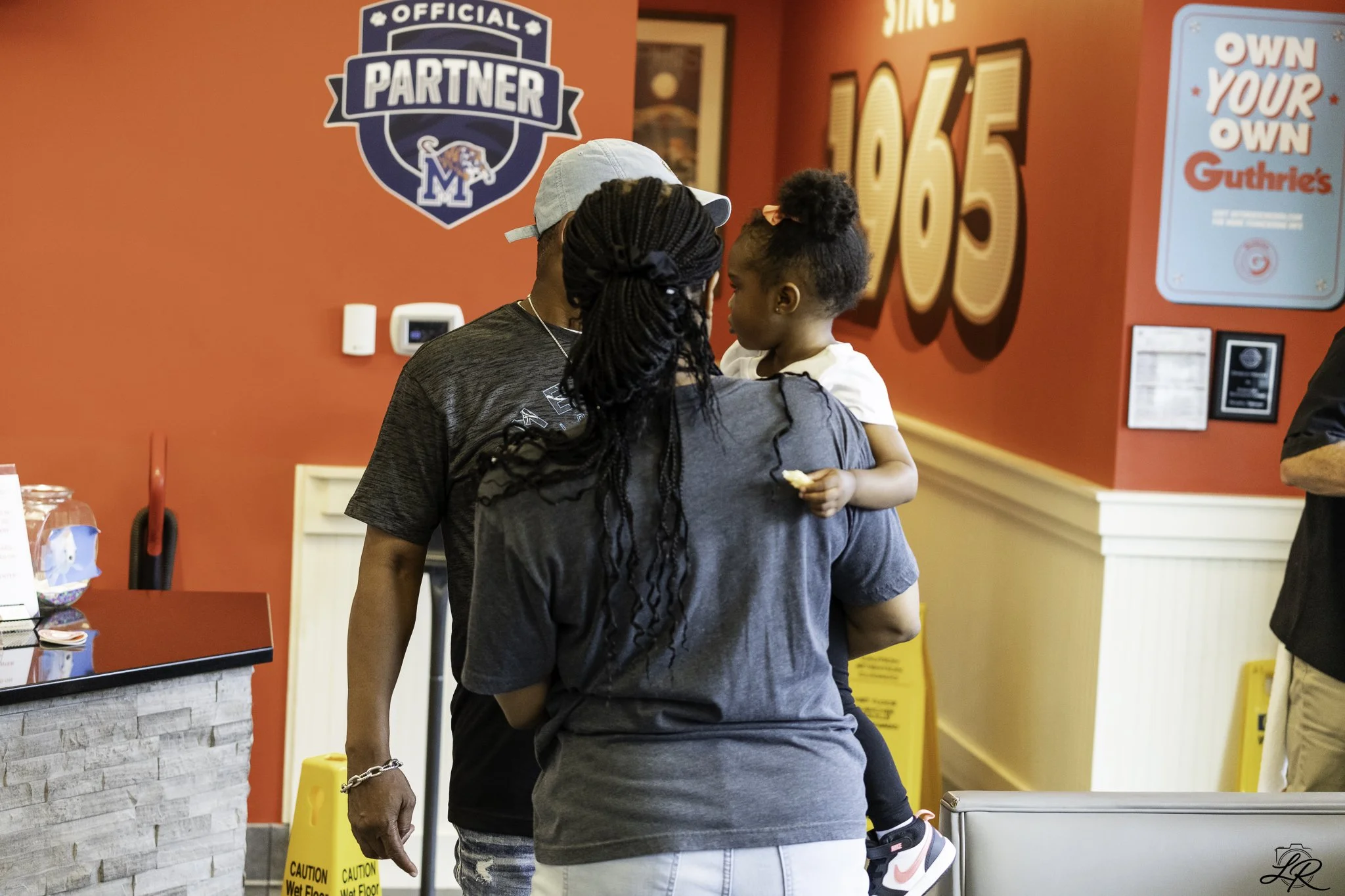 A man and a young girl share a close moment in a fast-food restaurant. The man has dark skin and braided hair, wearing a grey T-shirt and a light-colored cap. The girl has dark skin and curly hair tied with a pink bow, wearing a grey T-shirt and blac