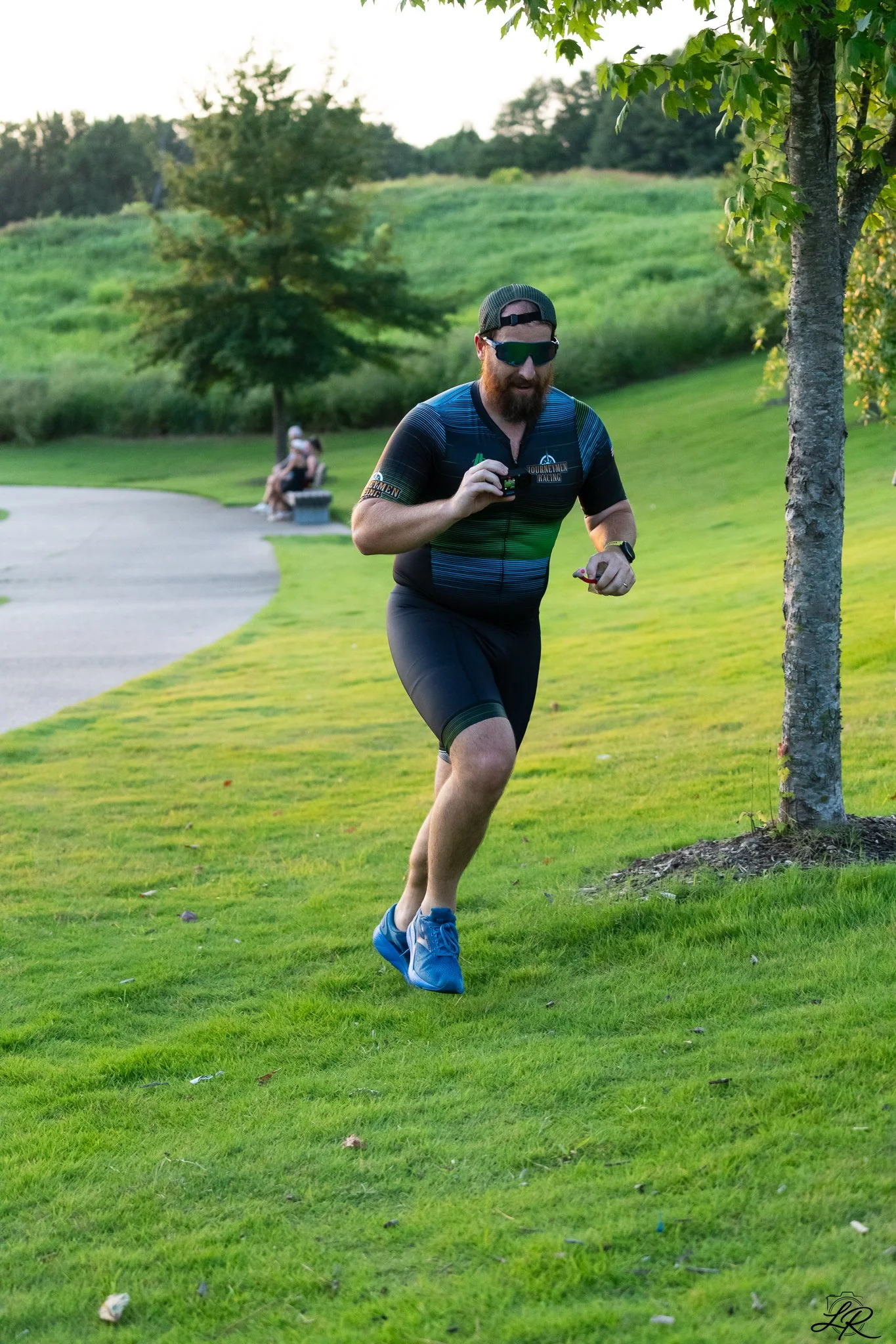 A man with a beard, sunglasses, and a green cap running on a grassy area near a tree in a park during daylight.