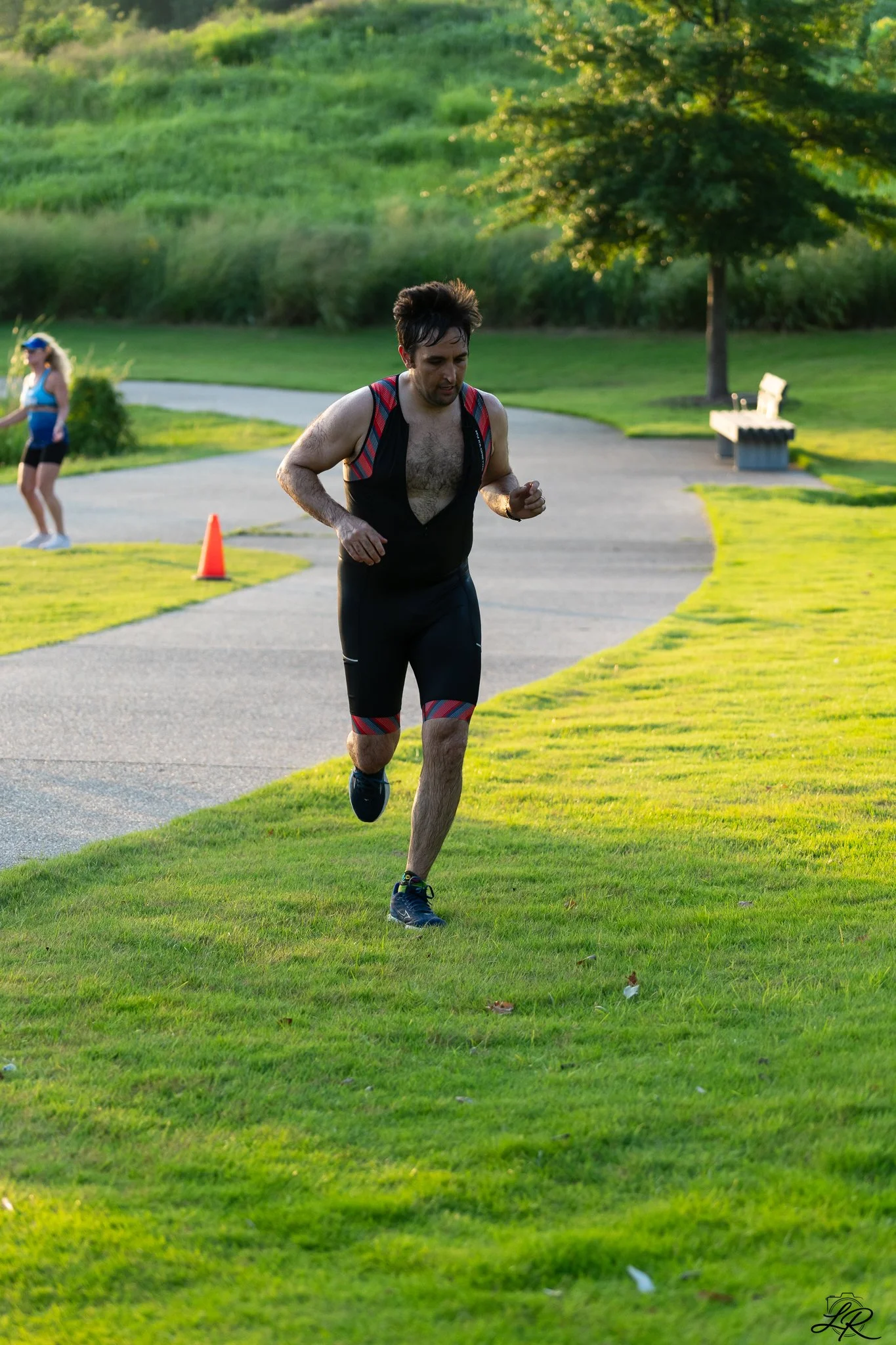 A man running on a grassy park path during the daytime, with another person in the background, a tree, and green hills.
