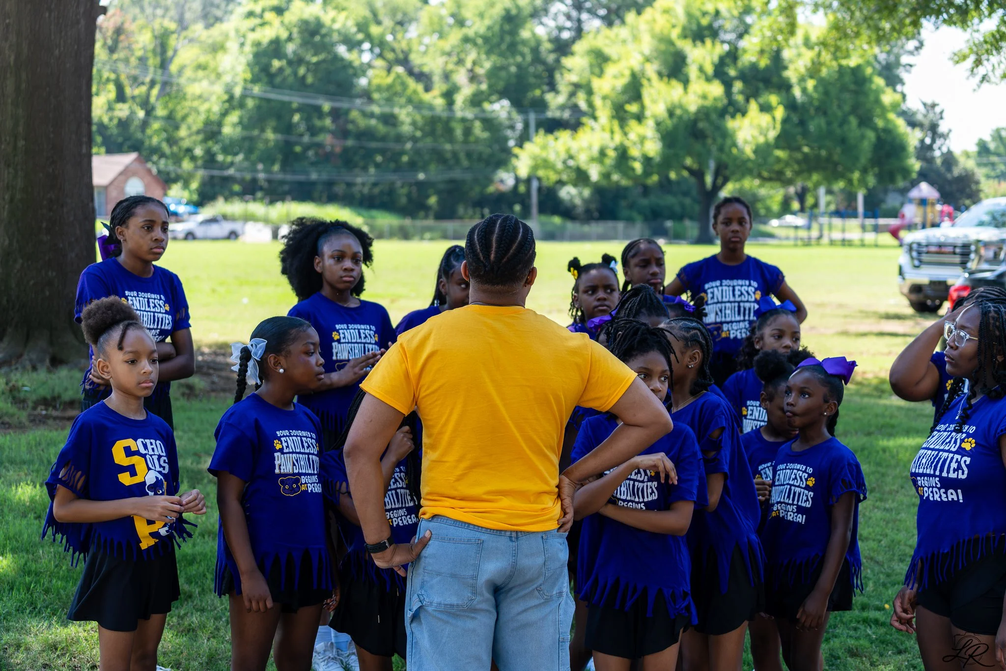 A group of young girls wearing matching blue shirts with yellow text and a bear graphic, gathered outdoors in a park on a sunny day, listening to an adult who is speaking to them.