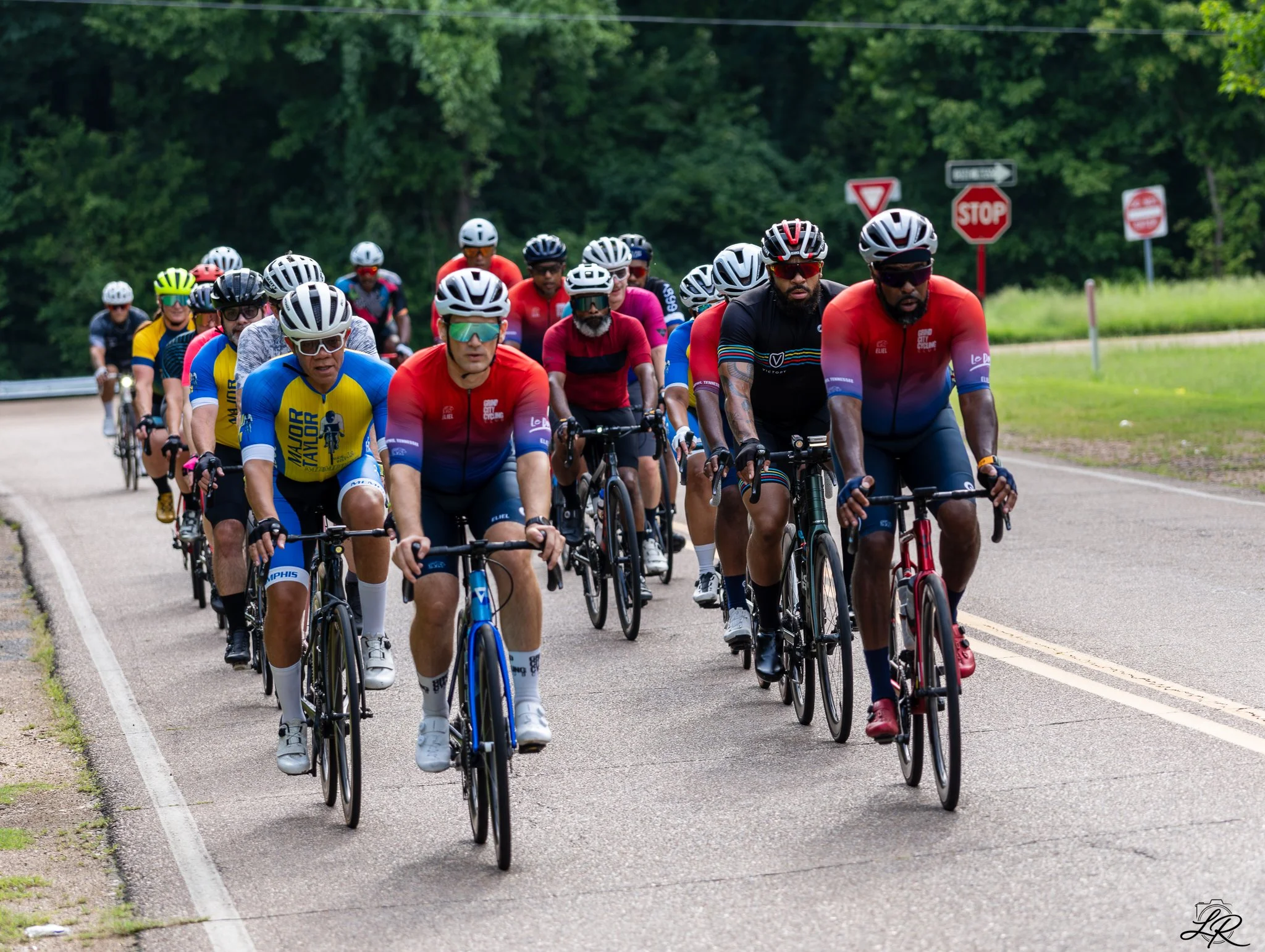 A large group of cyclists riding on a paved road with lush green trees in the background, some stop signs and a street sign visible.