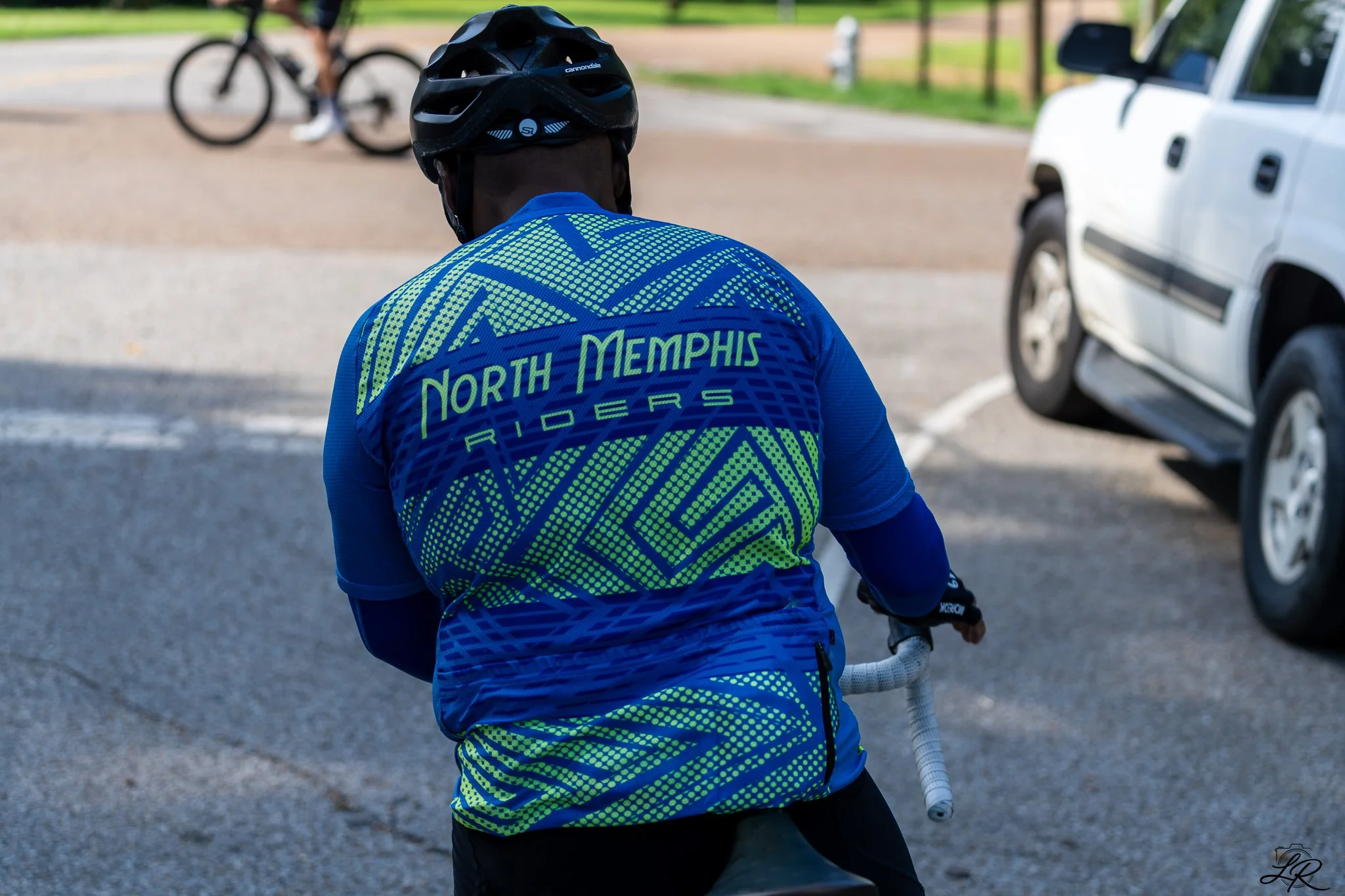 Person wearing a blue and yellow North Memphis Riders cycling jersey and helmet, sitting on a bike with a white handlebar, with a cyclist and cars in the background.