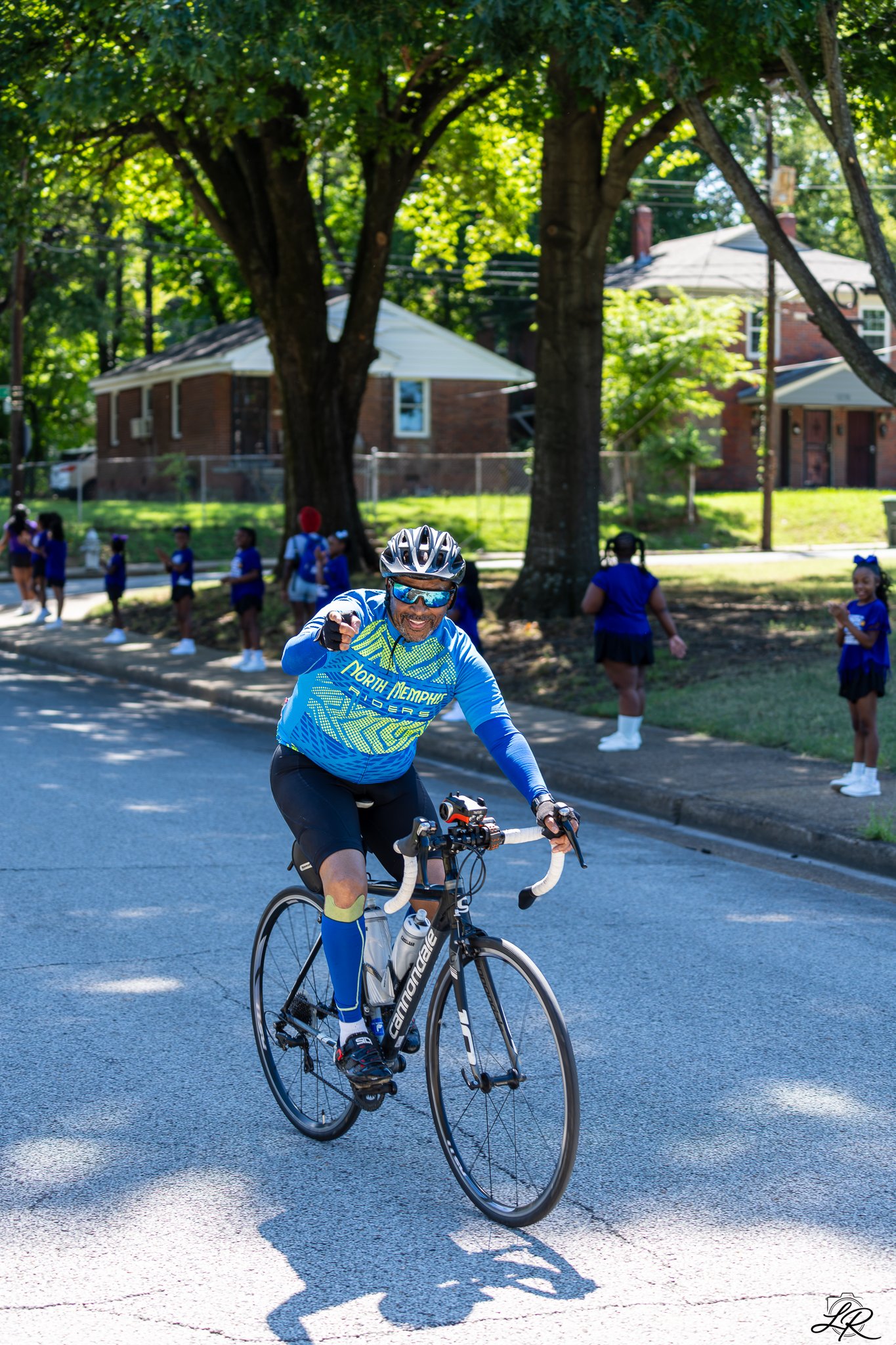 A man riding a black Cannondale road bike on a street with shadows from trees, wearing a blue North Memphis cycling jersey, blue cycling shorts, a helmet, sunglasses, and gloves. In the background, children in blue uniforms are standing near trees an