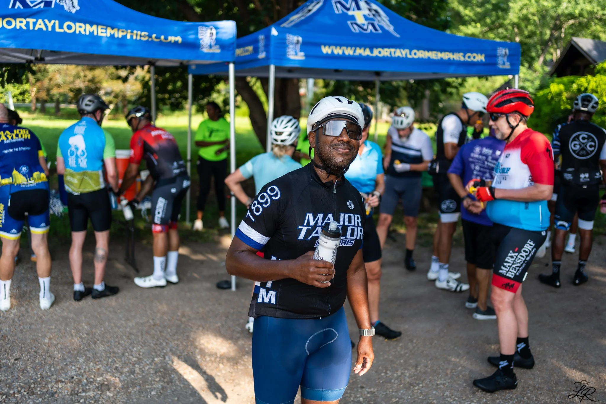 A group of cyclists in sports gear and helmets gathered under a large blue tent with 'Major Taylor' branding and website, some talking, others drinking water, in a park setting.