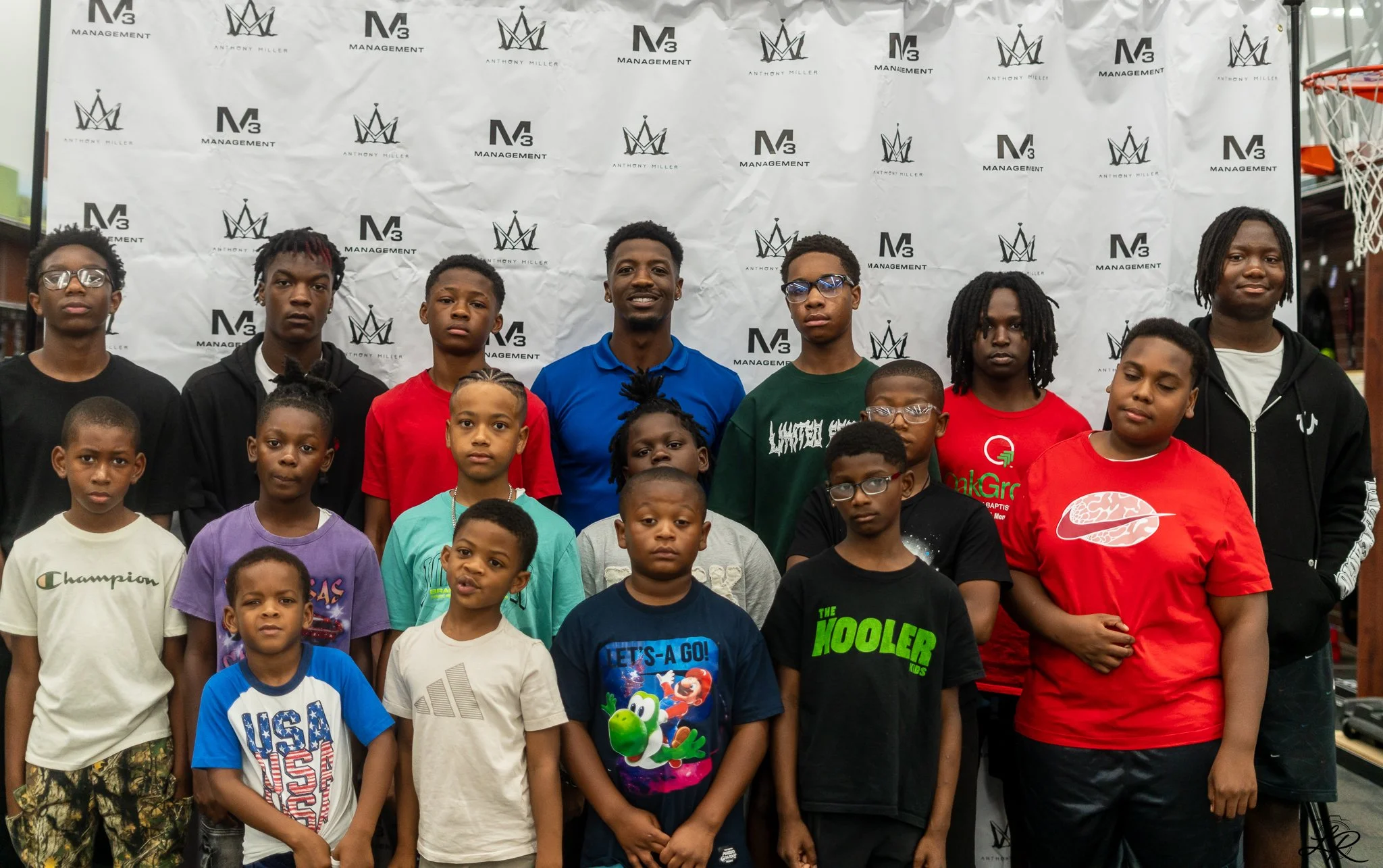 A group of children and teenagers standing in front of a step-and-repeat banner with logos for 'Anthony Miller' and 'M3 Management,' posing for a photo indoors near a basketball hoop.