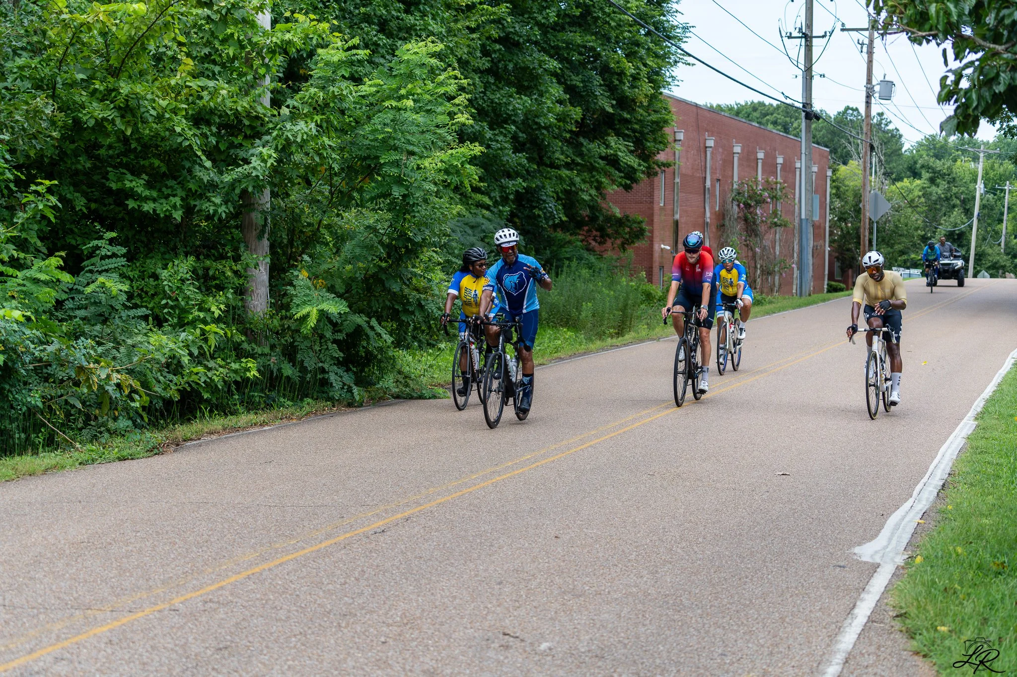 Group of cyclists riding on a rural road surrounded by green trees and a brick building, with utility poles and power lines overhead.
