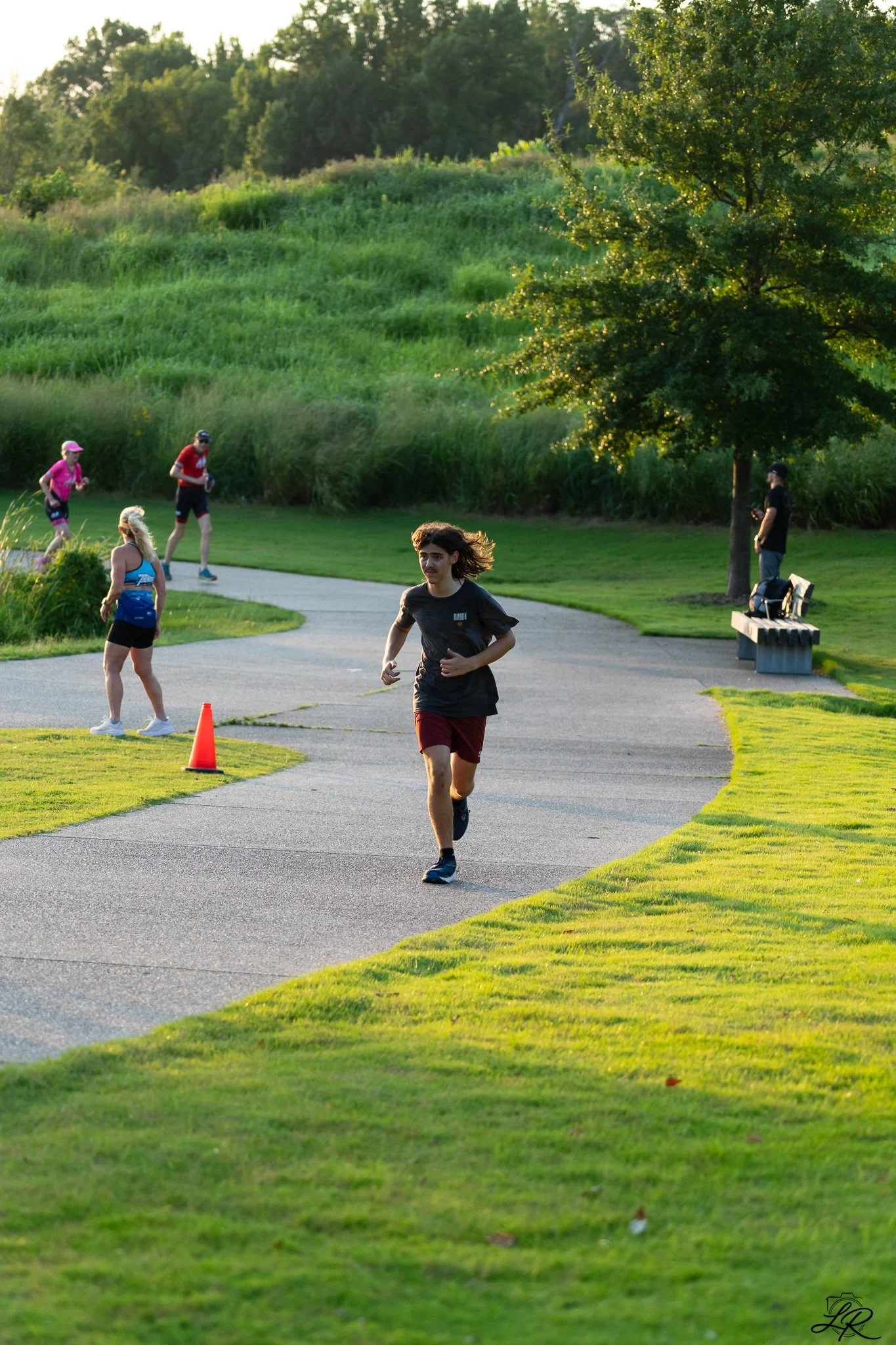 A man running on a paved path in a park during late afternoon or early evening, with two women walking or jogging behind him, a person standing near a tree holding a phone, a wooden bench nearby, green trees, a grassy area, and a hilly landscape in t