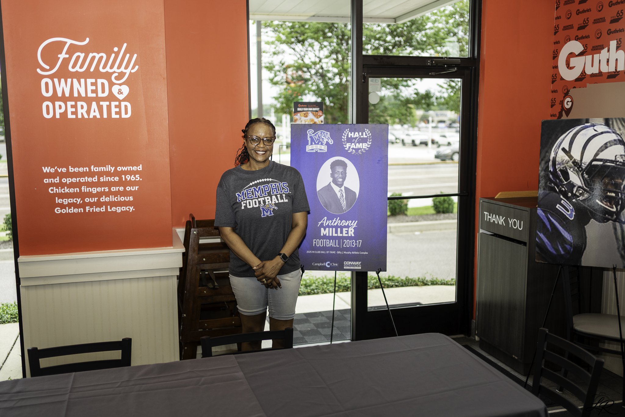 A woman smiling and standing next to a blue poster with a photo of an athlete named Anthony Miller, in a room with large windows, red walls with Memphis branding, and a table with a gray tablecloth.
