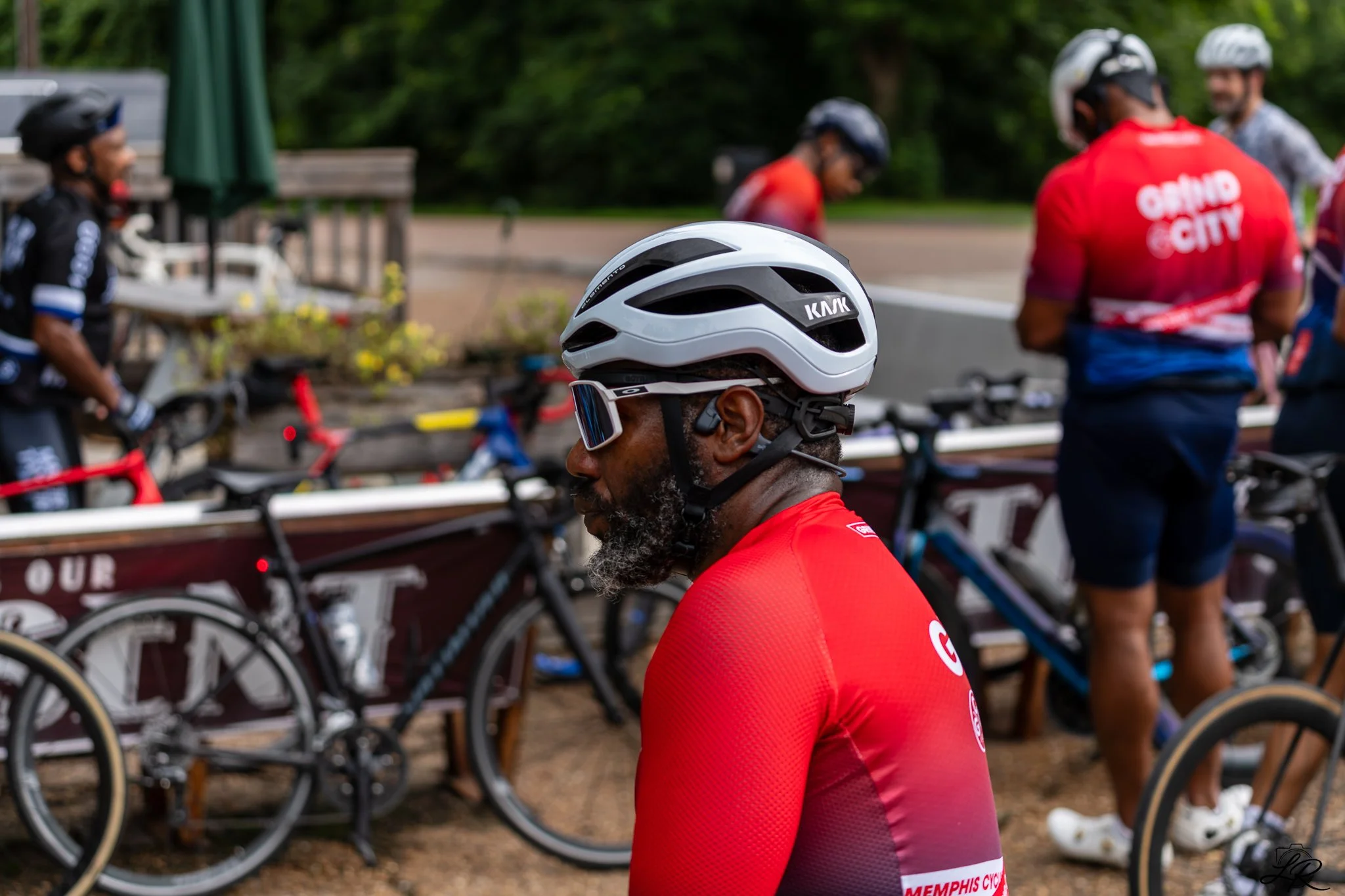 A man wearing a white bicycle helmet, sunglasses, and a red cycling jersey stands in front of bicycles and other cyclists in a park.