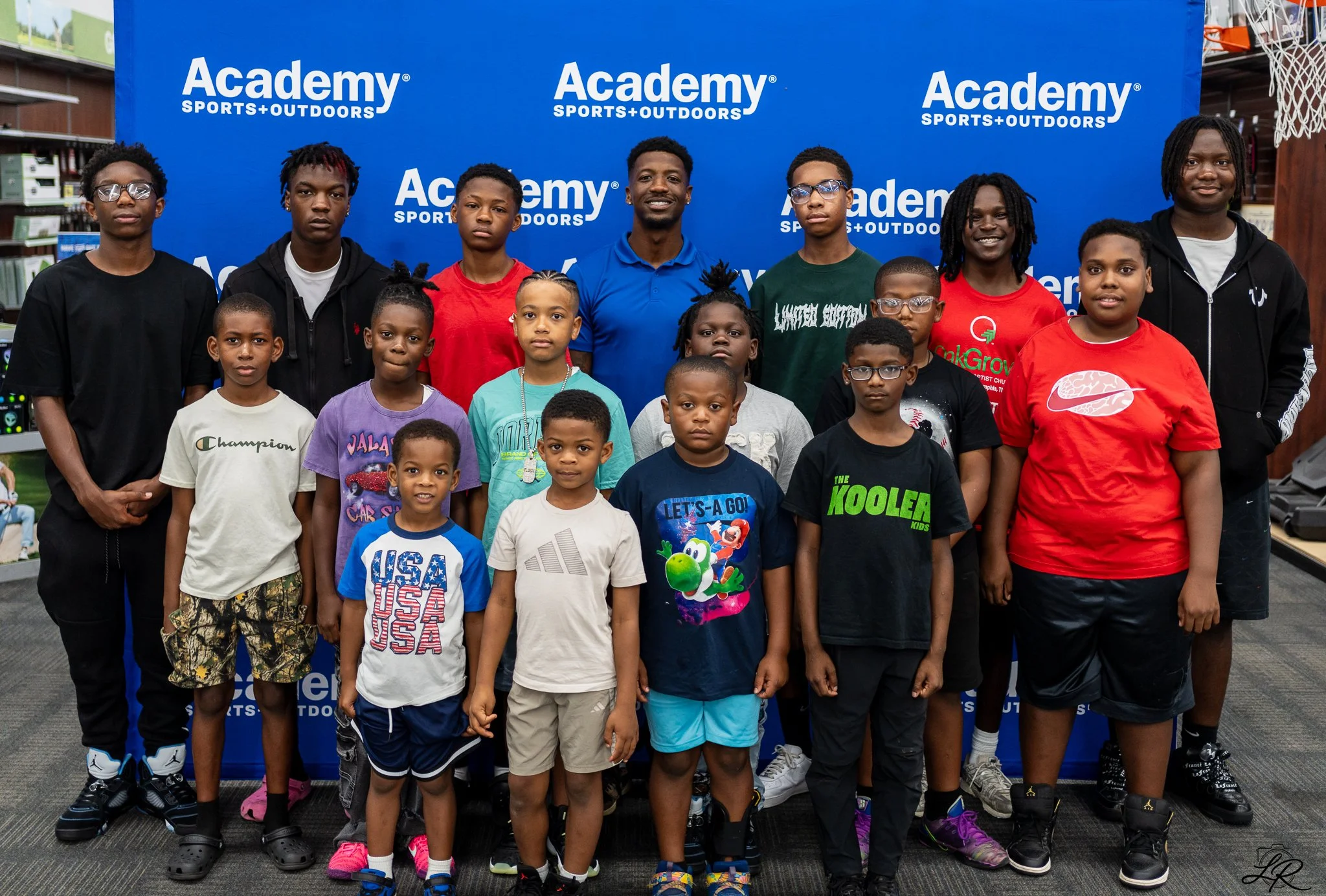 Group of children and teenagers standing in front of a blue backdrop with 'Academy Sports+Outdoors' logo, indoors at a store.