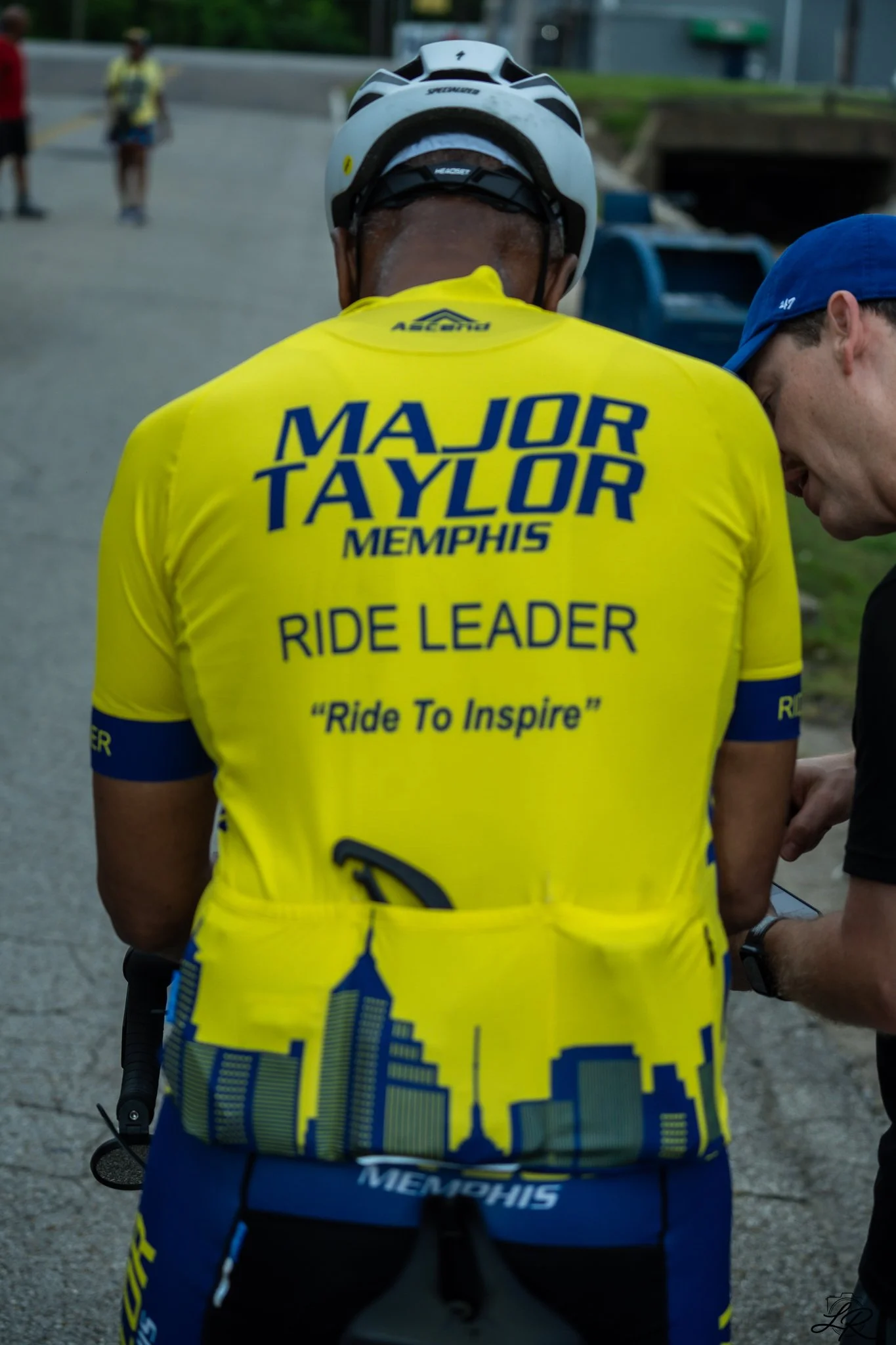 A man wearing a bright yellow cycling jersey with 'Major Taylor Memphis' and 'Ride to Inspire' written on the back, and a white helmet. He is standing on a street with a person in a blue cap beside him, and other cyclists and pedestrians in the backg