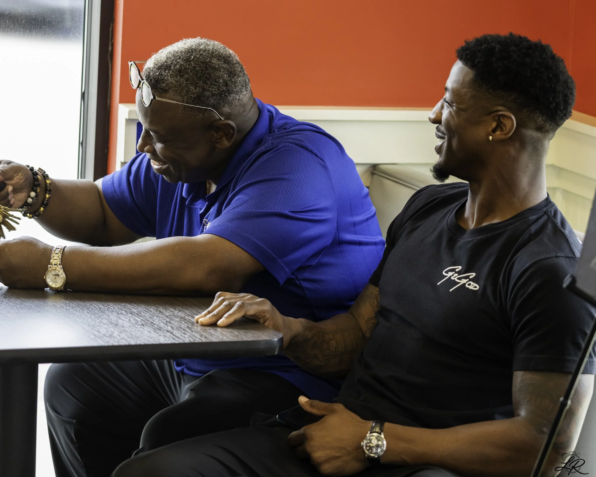 Two men sitting side by side in a room with a red wall, engaged in a conversation. The man on the left is older with gray hair, glasses on his head, wearing a blue collared shirt, a gold watch, and beaded bracelets. The man on the right is younger, w