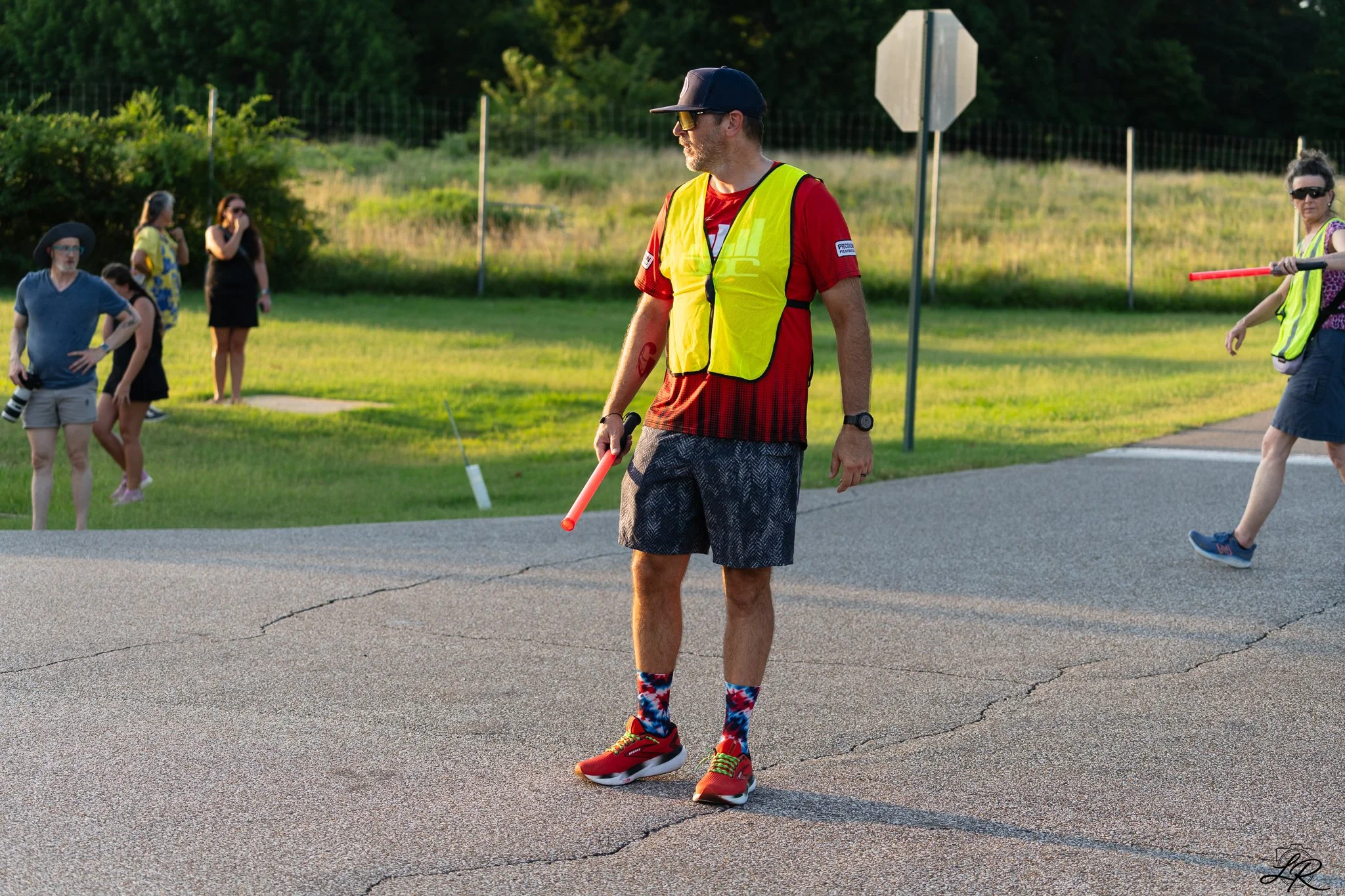 A man in a red shirt, black shorts, and colorful socks holds a traffic baton on a road, surrounded by women in casual clothing and high-visibility vests, with greenery and a stop sign in the background.