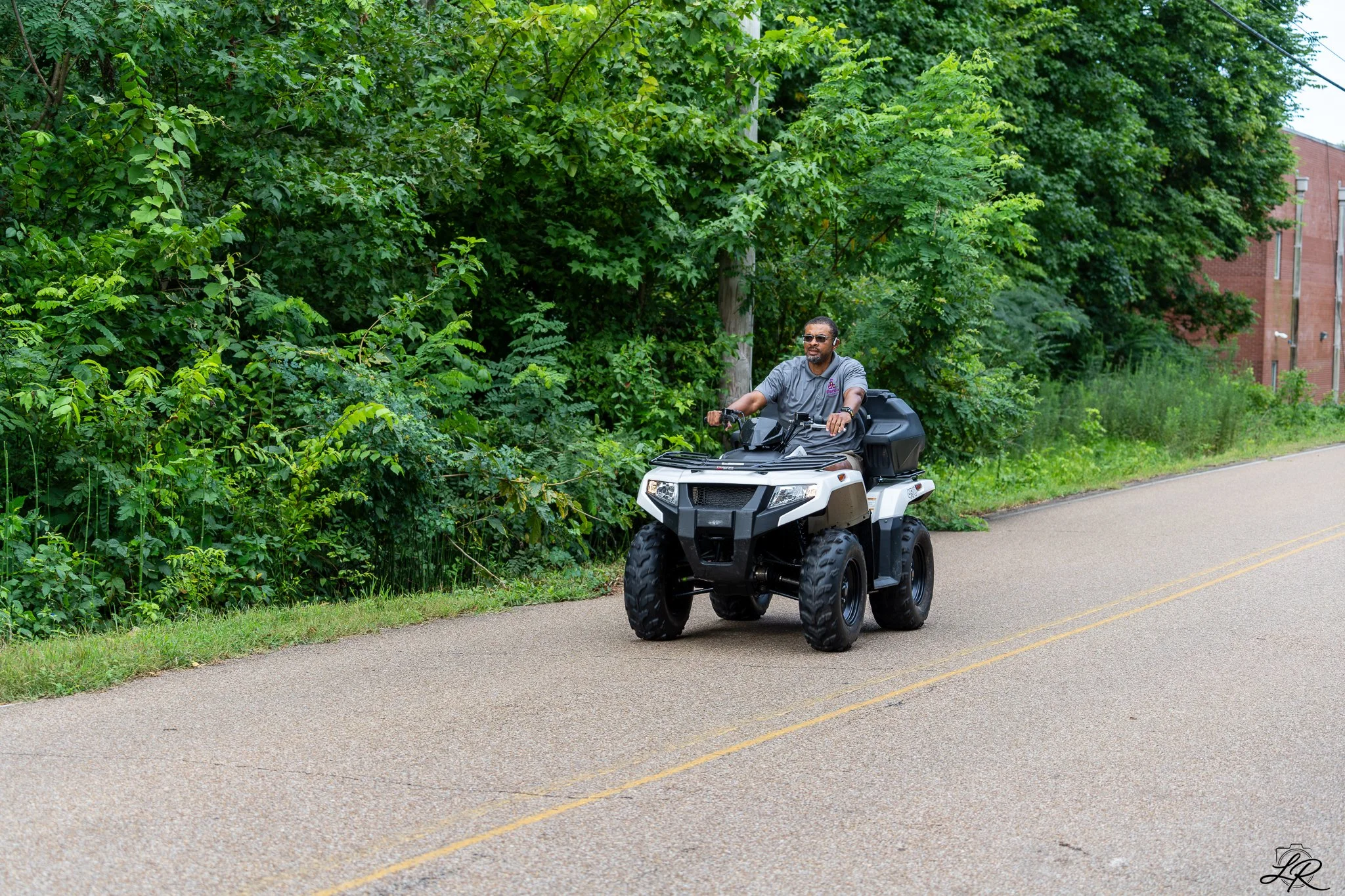 A man riding an all-terrain vehicle (ATV) on a paved road beside a dense, green wooded area.